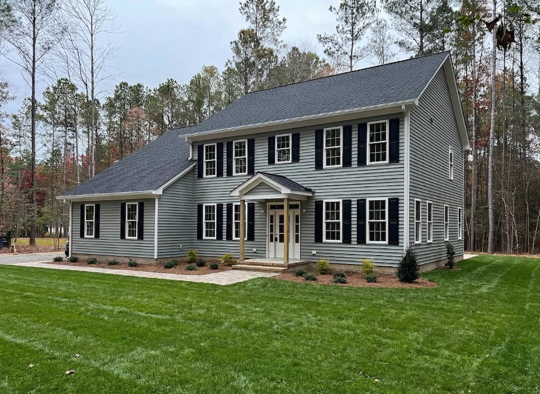 A two-story house with gray exterior, black shutters, and a front porch with two pillars, situated on a well-maintained lawn with small shrubs, surrounded by trees in the background.
