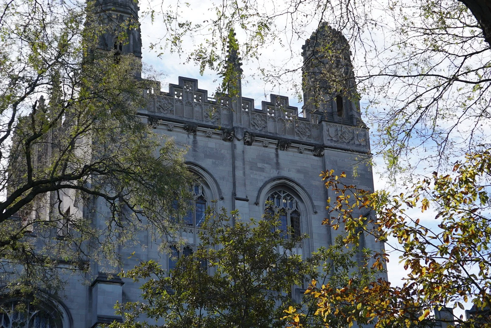 A view of a historic stone church with Gothic windows and tall towers, partially obscured by trees with green and yellow leaves, under a sky with clouds.