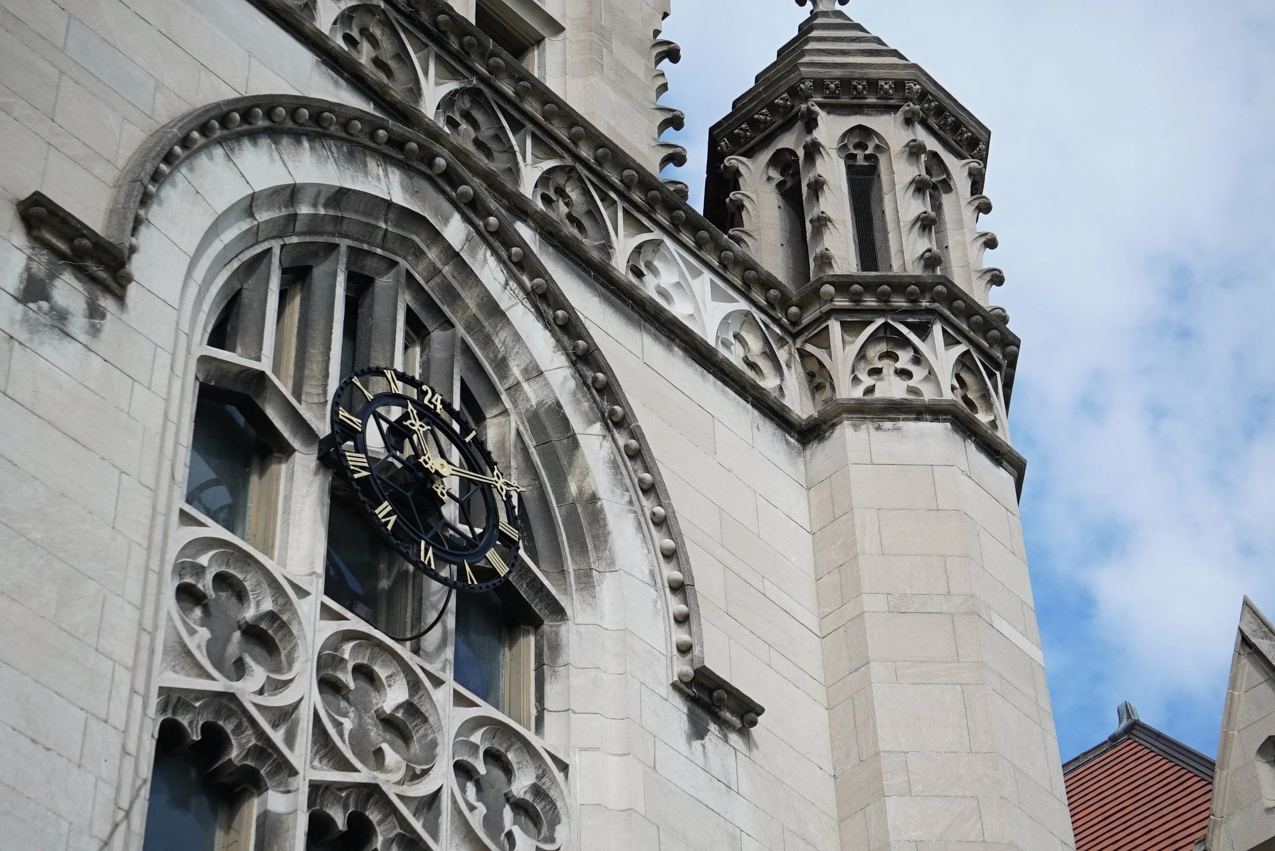 Close-up of a historic stone building with Gothic architectural details, including ornate carvings and a clock with Roman numerals, under a partly cloudy sky.