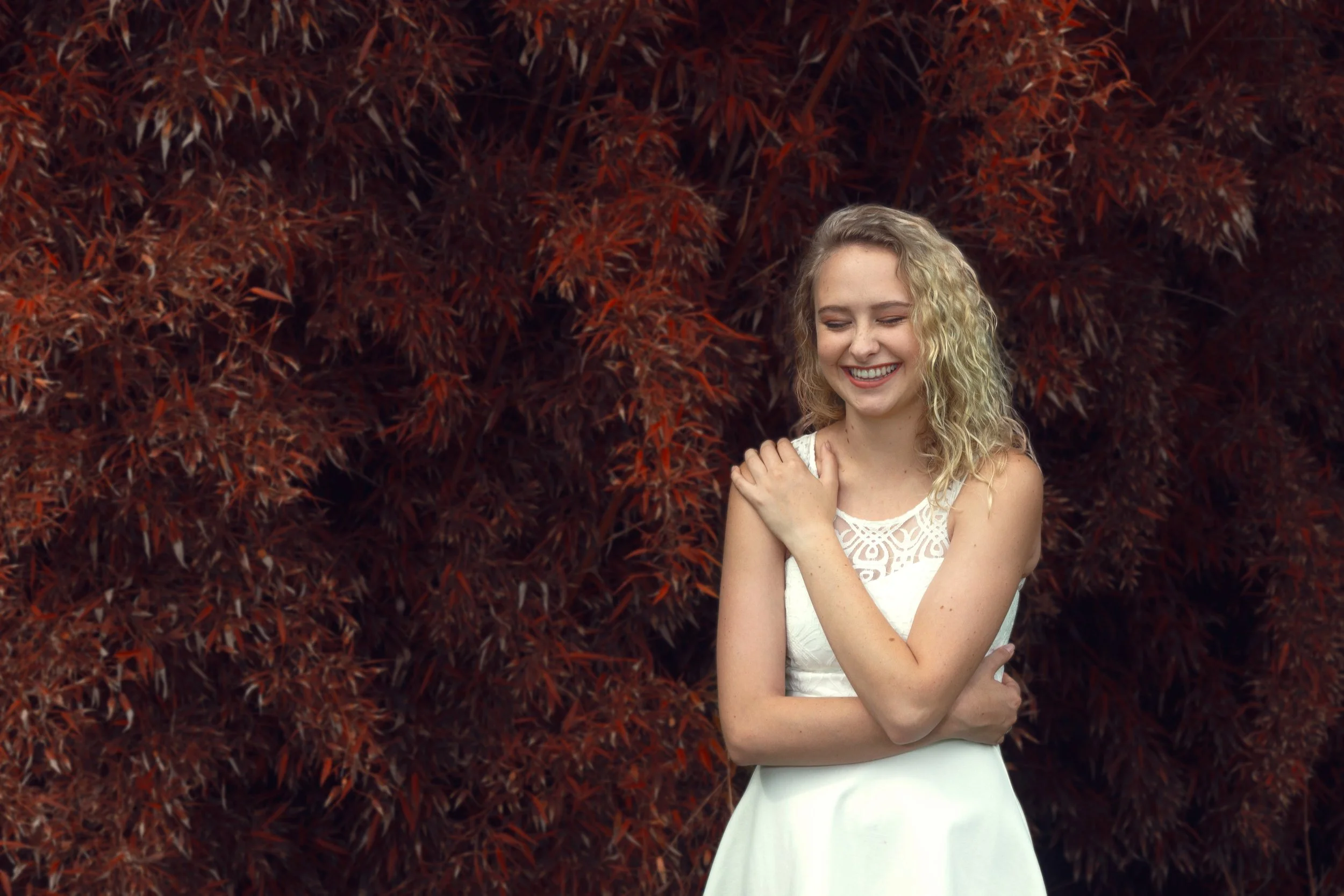 A woman with blonde curly hair smiling and standing in front of red autumn leaves. She wears a white sleeveless dress and has her arm crossed over her chest, holding her shoulder.