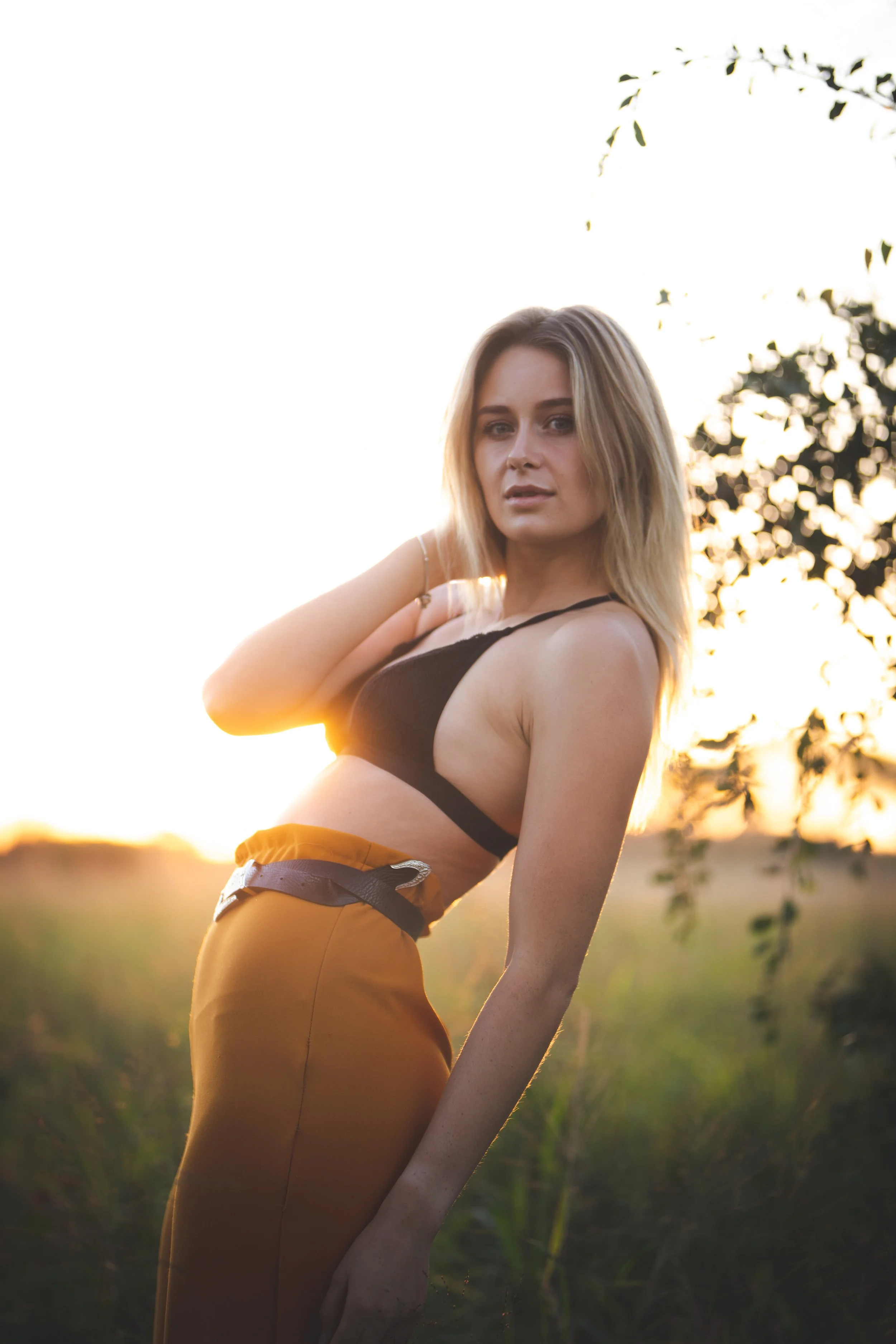 A young woman with blonde hair posing outdoors during sunset, wearing a black crop top and orange high-waisted pants, with one hand behind her head and the other hanging by her side.