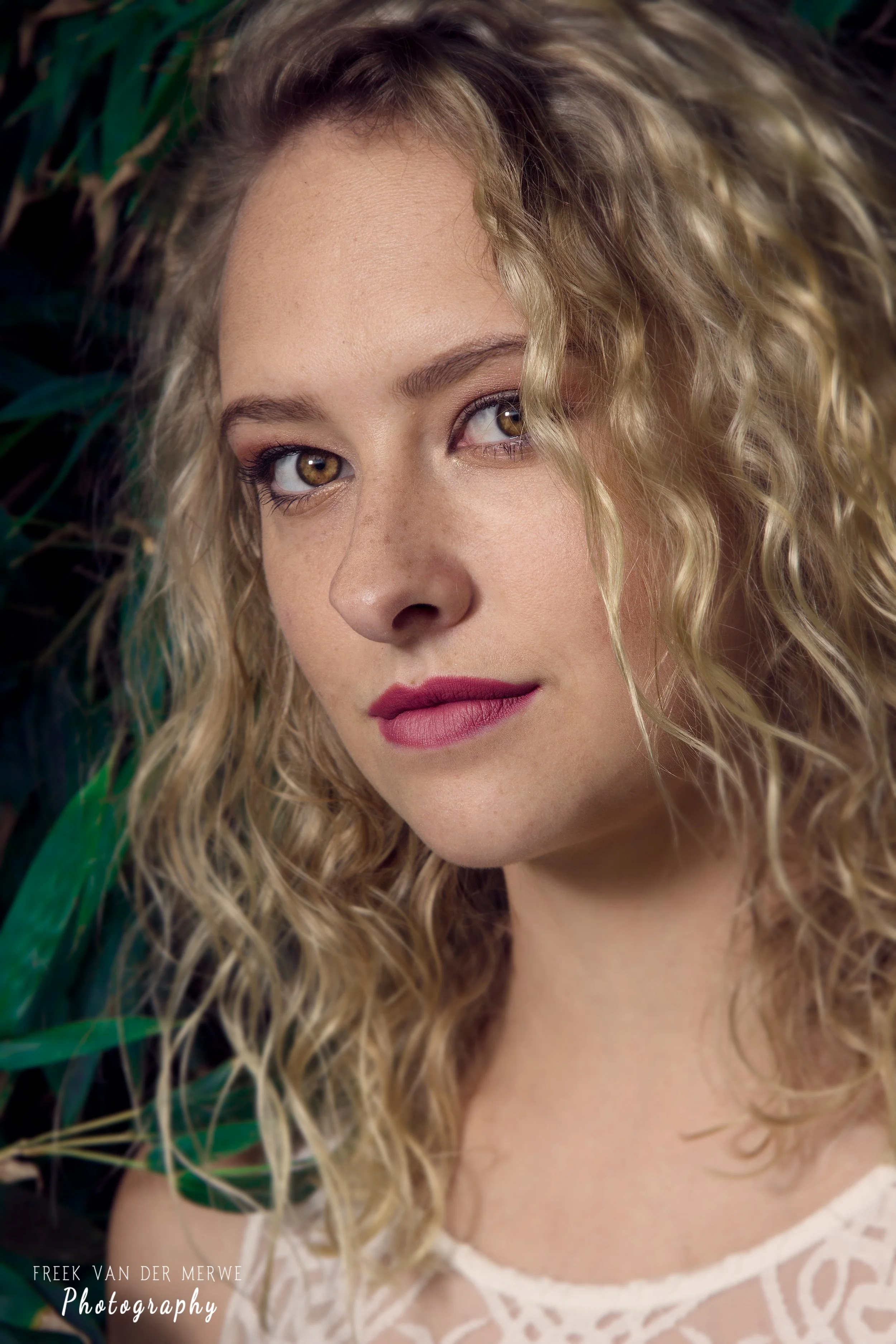 Close-up portrait of a woman with curly blonde hair, fair skin, and brown eyes, looking at the camera, with tropical leaves in the background.