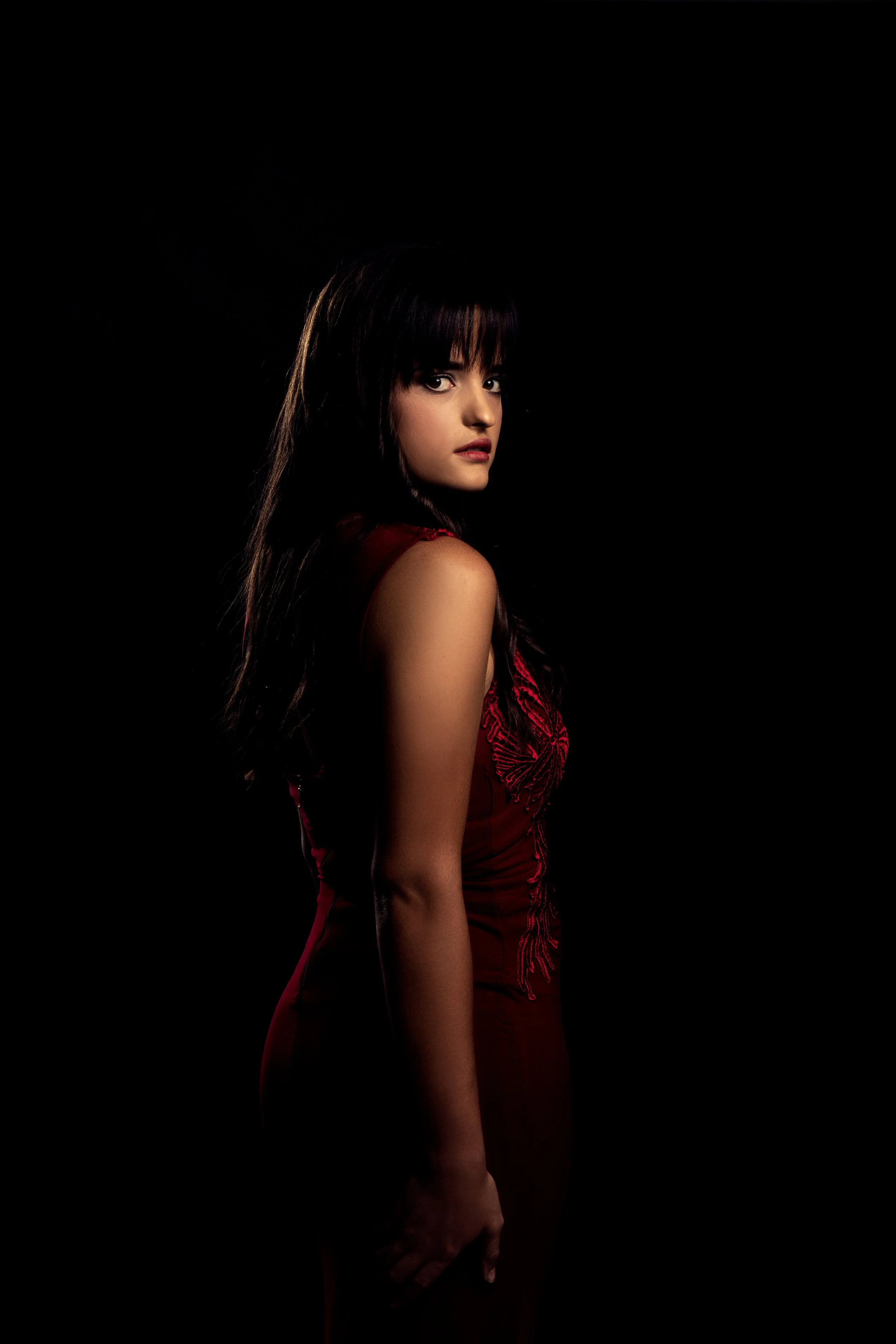 A woman with dark hair wearing a red dress standing against a black background.