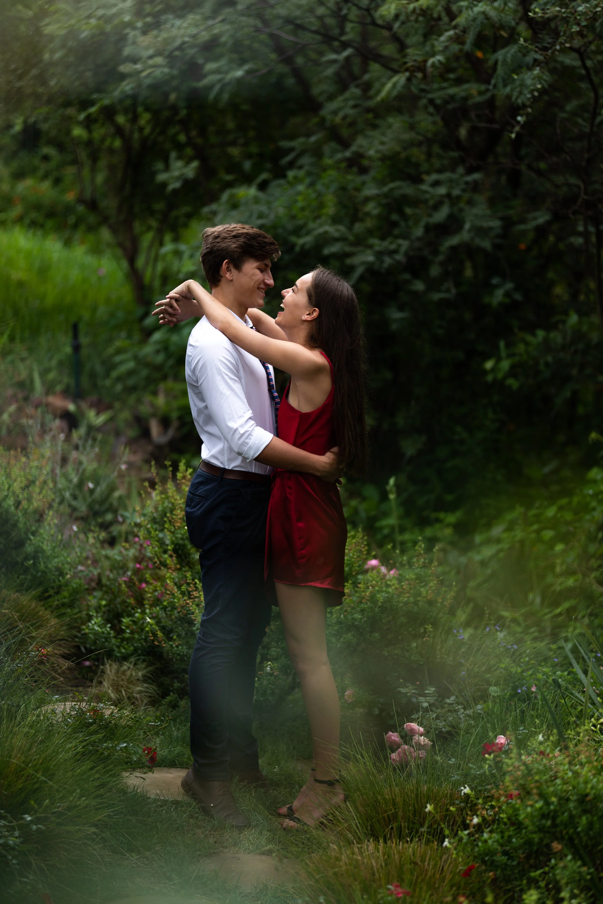 A couple embracing in a lush green garden, smiling at each other, with trees and flowers surrounding them.