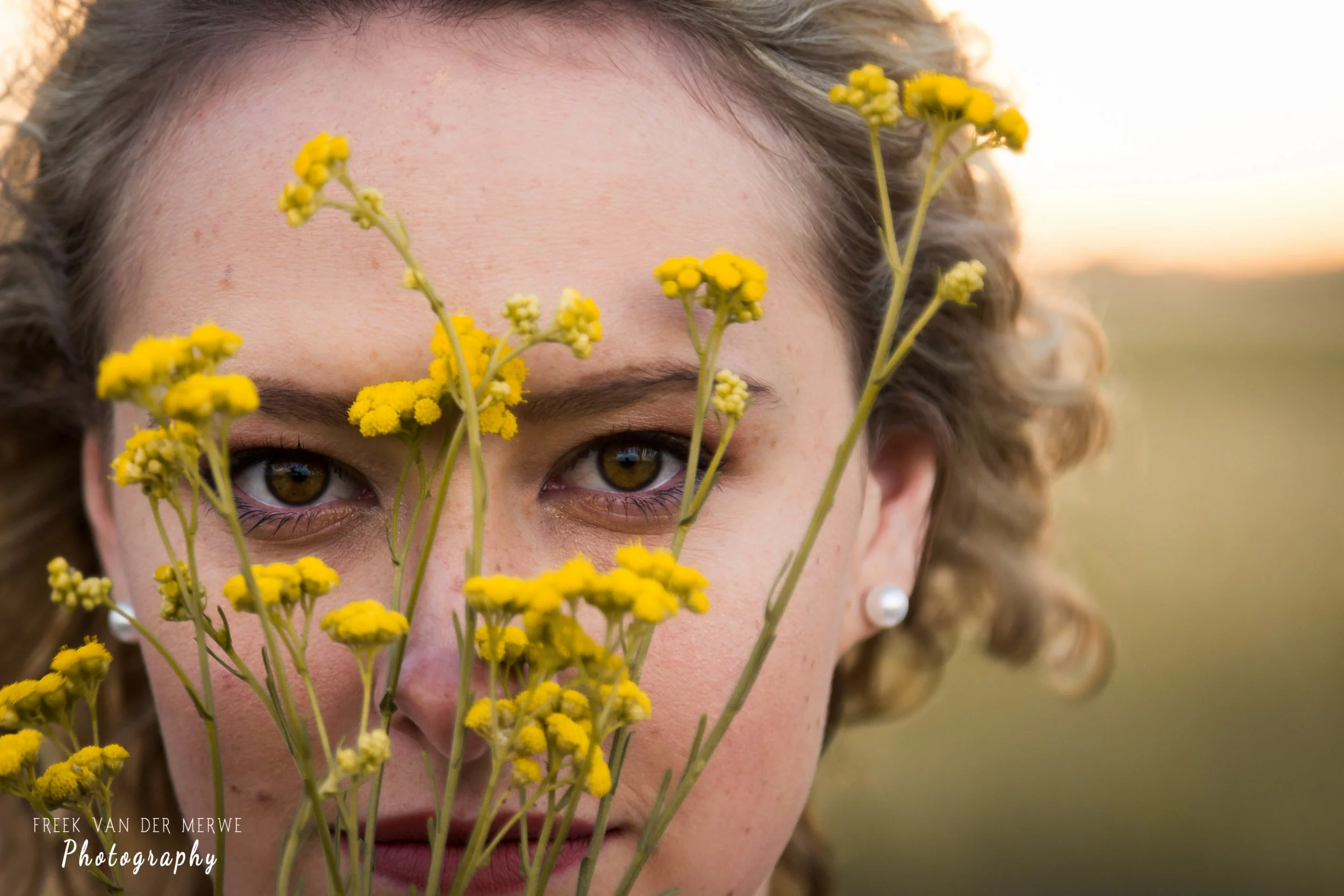 Close-up of a woman with curly hair and brown eyes, holding yellow flowers close to her face, with a blurred outdoor background at sunset.