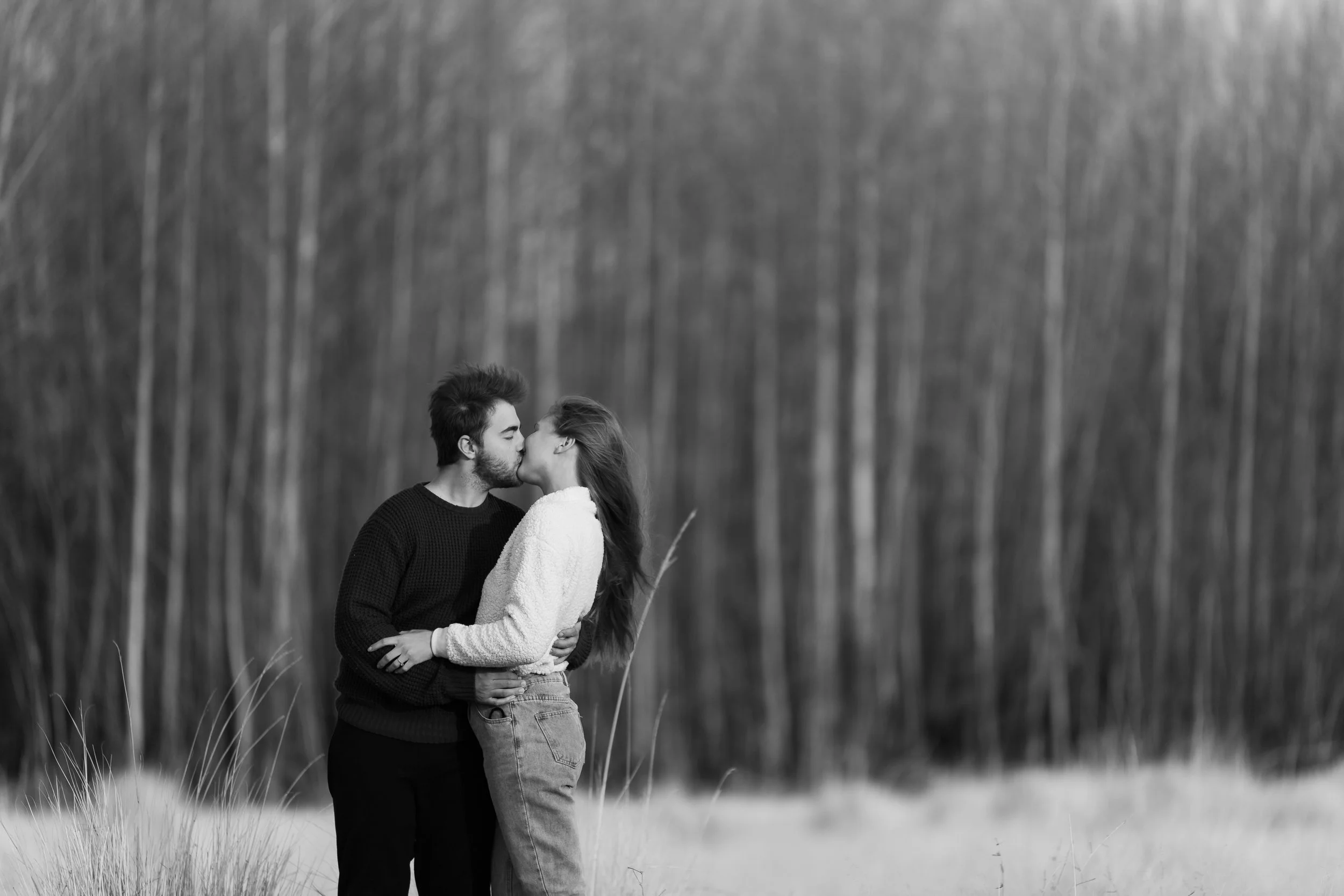 A black and white photo of a young couple kissing outdoors with blurred trees in the background.
