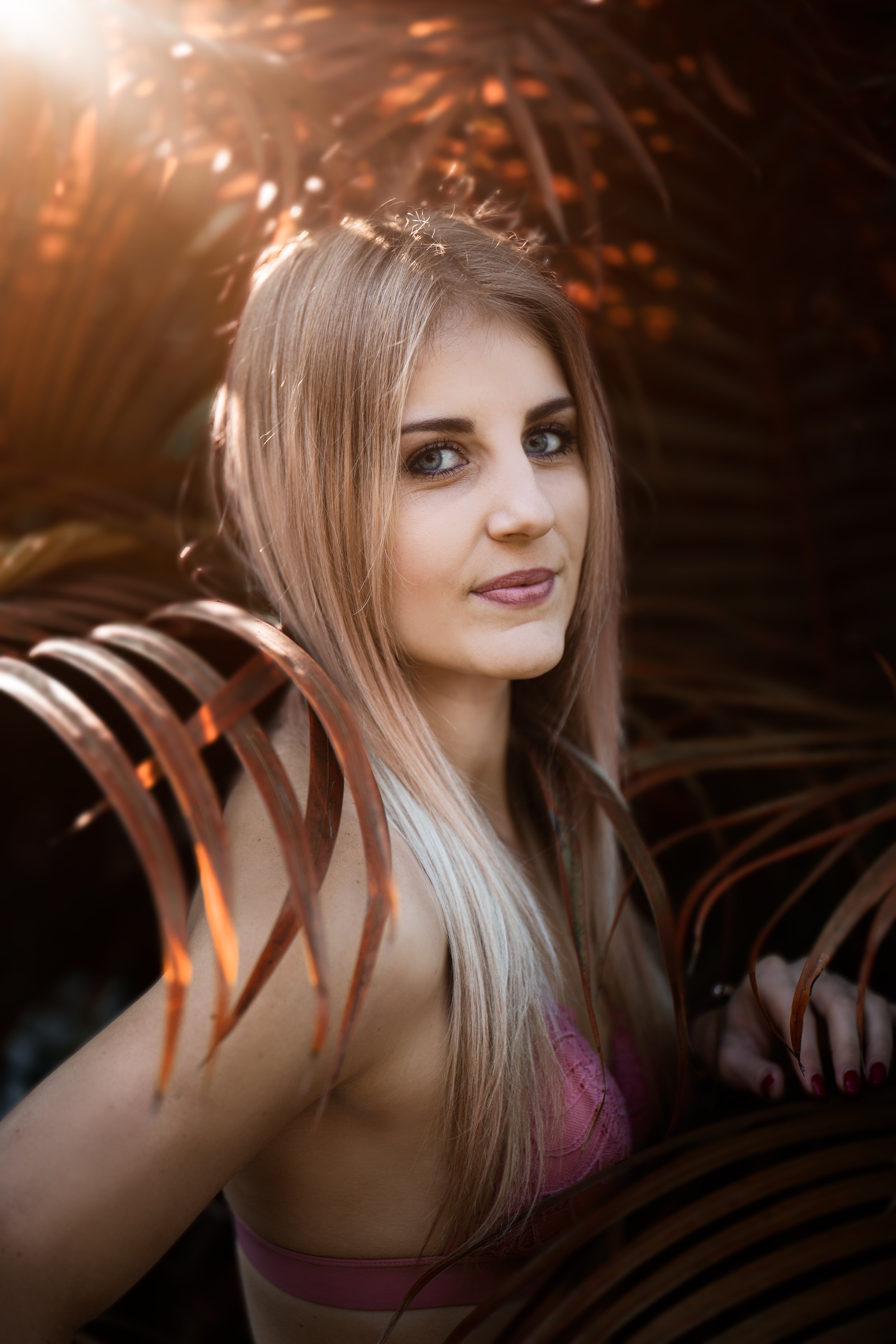 A woman with long blonde hair and blue eyes looking at the camera, surrounded by dark brown palm leaves with sunlight filtering through in the background.