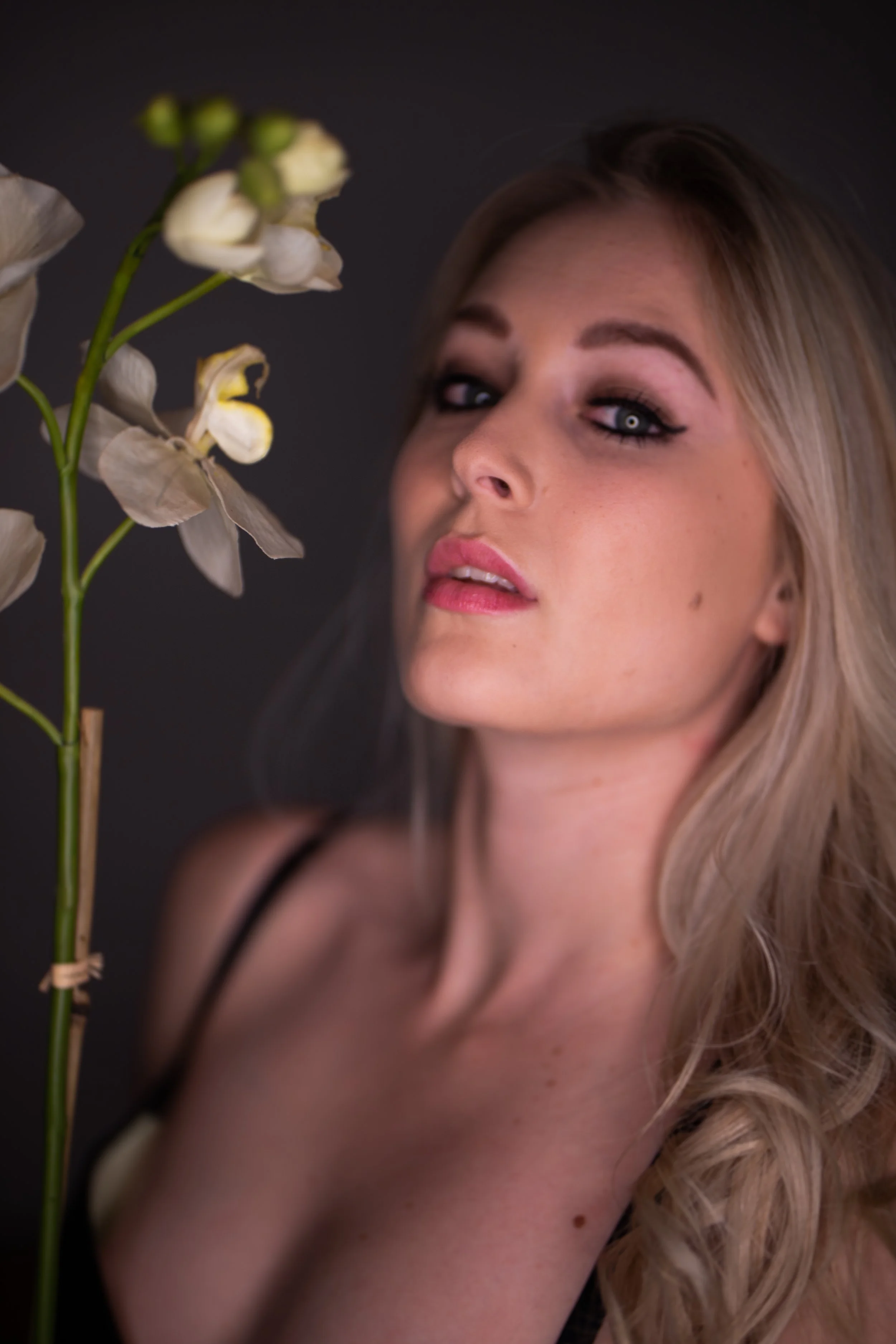 A woman with blonde hair and makeup gazes at a white flower on a dark background.