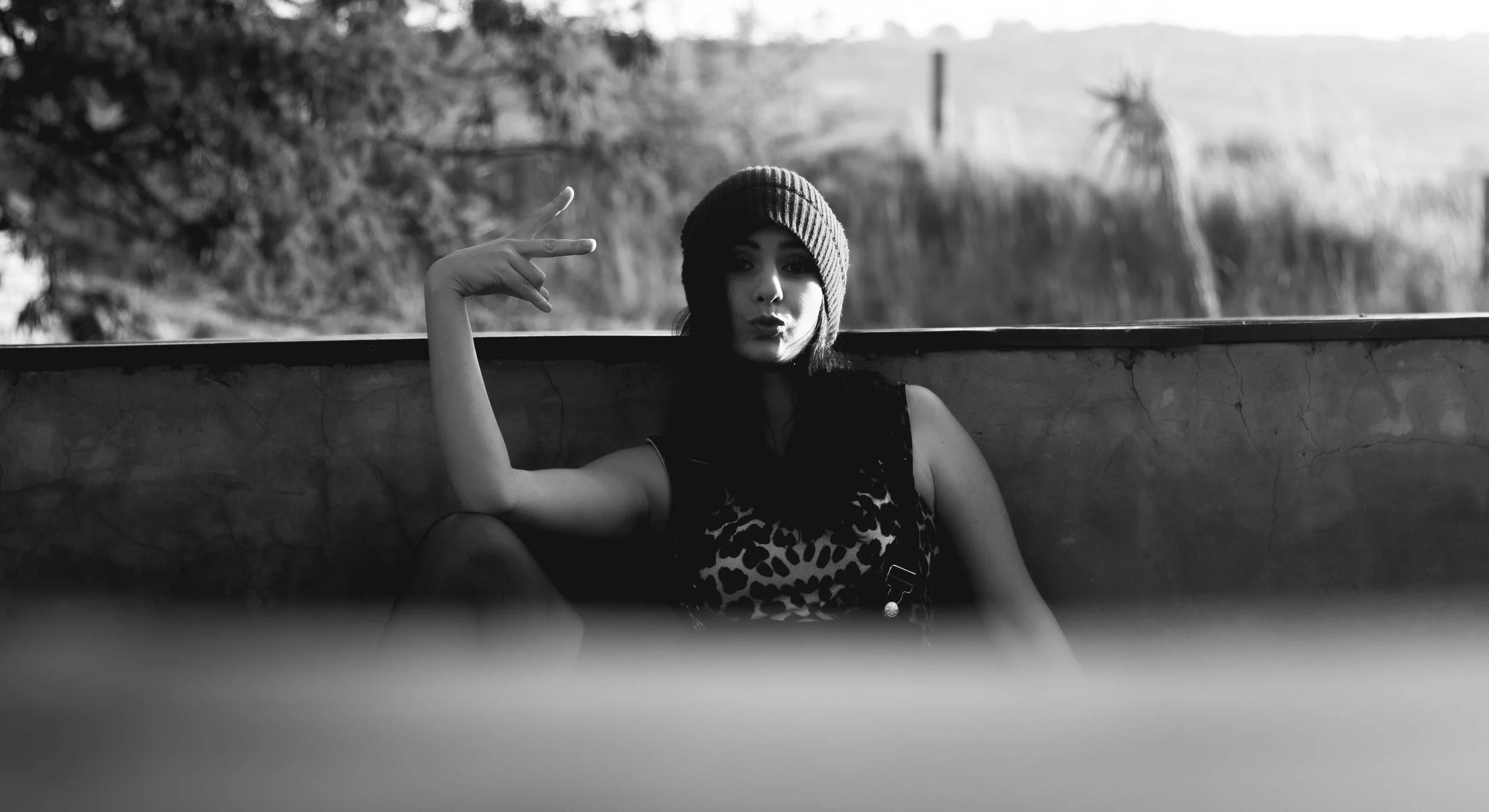 A young woman wearing a beanie and a leopard-print top, posing with her arm flexed and lips puckered outdoors in black and white.