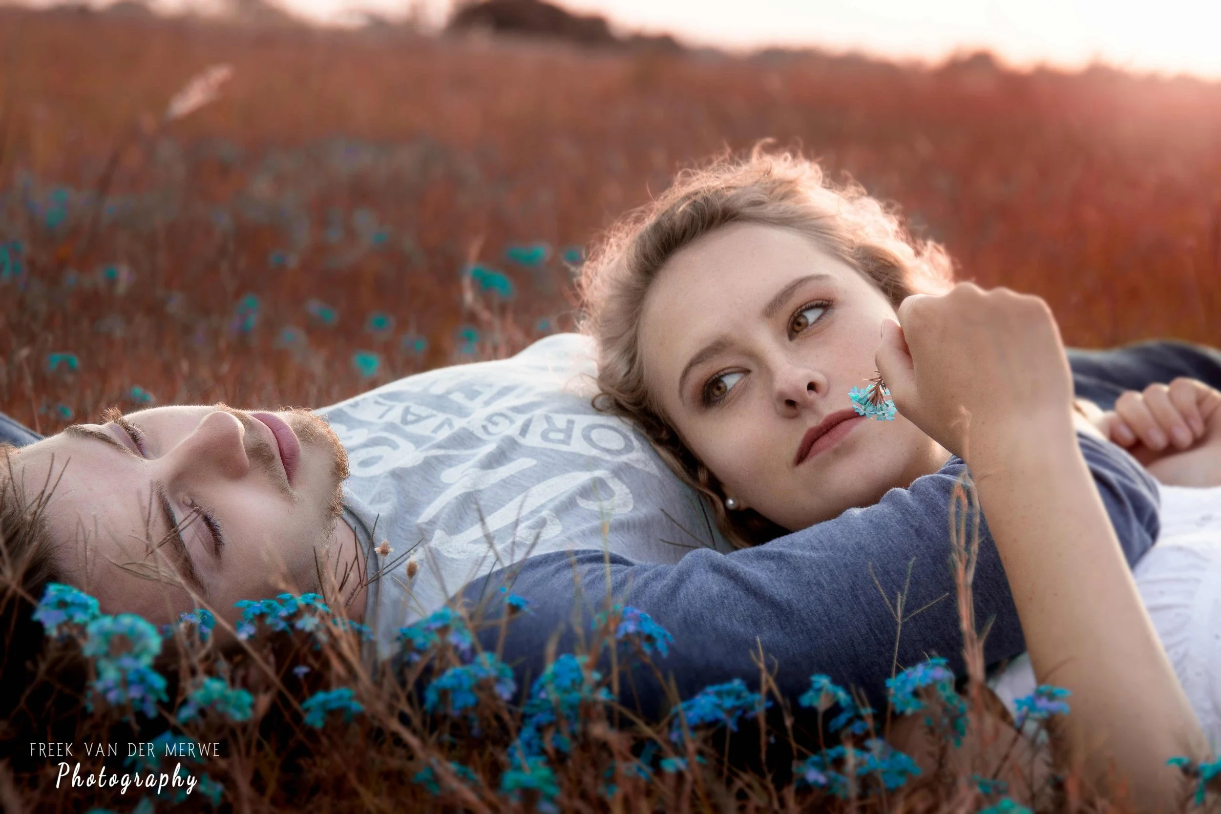 A young woman and man lying on the ground in a field of small blue flowers, with the woman resting her head on the man's arm and looking into the distance during sunset.
