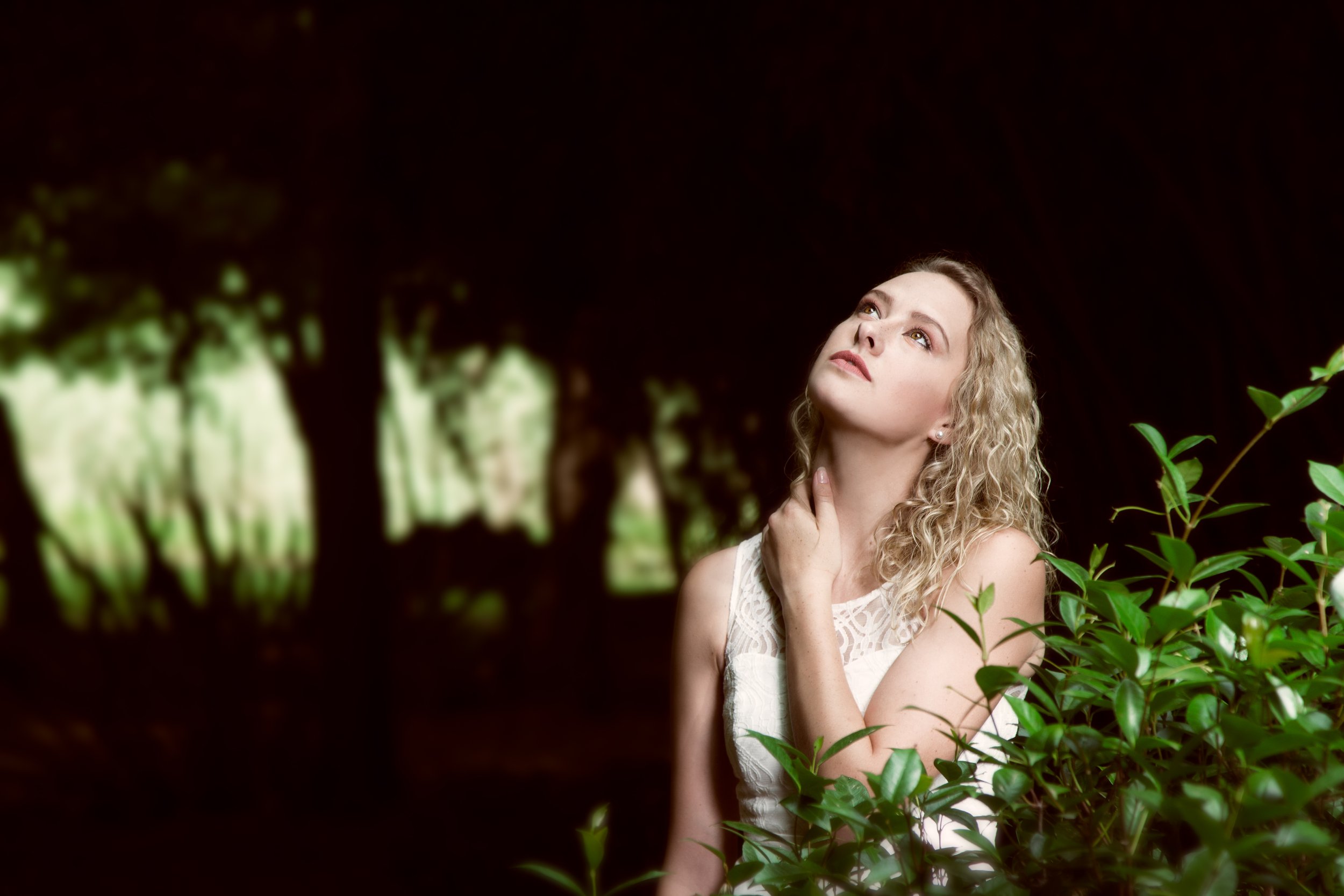 A young woman with curly blonde hair is posing outdoors among green bushes and trees, with one hand on her neck and looking up thoughtfully.