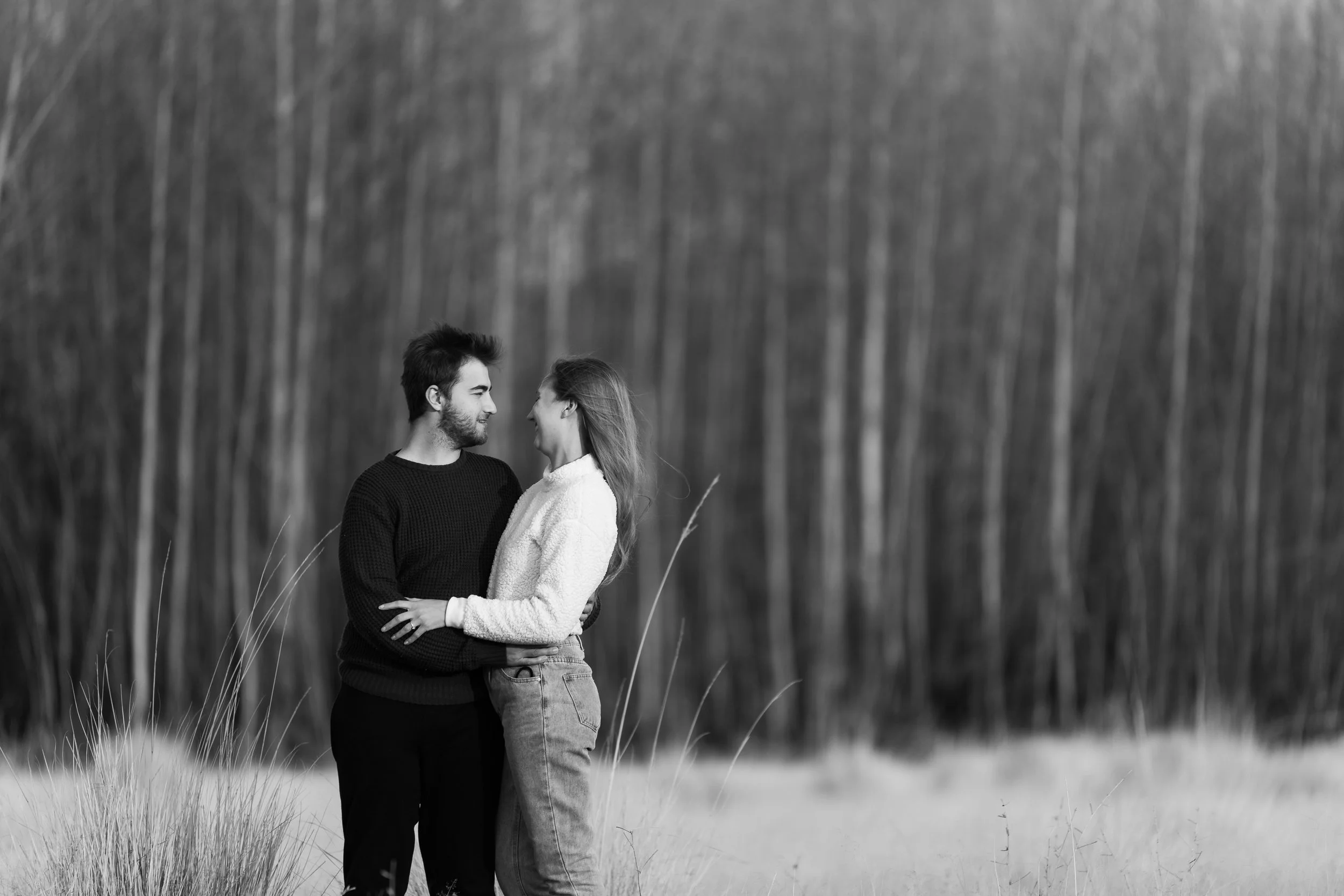 A black and white photo of a couple standing close together outdoors, holding each other and smiling at each other, with a forest in the background.