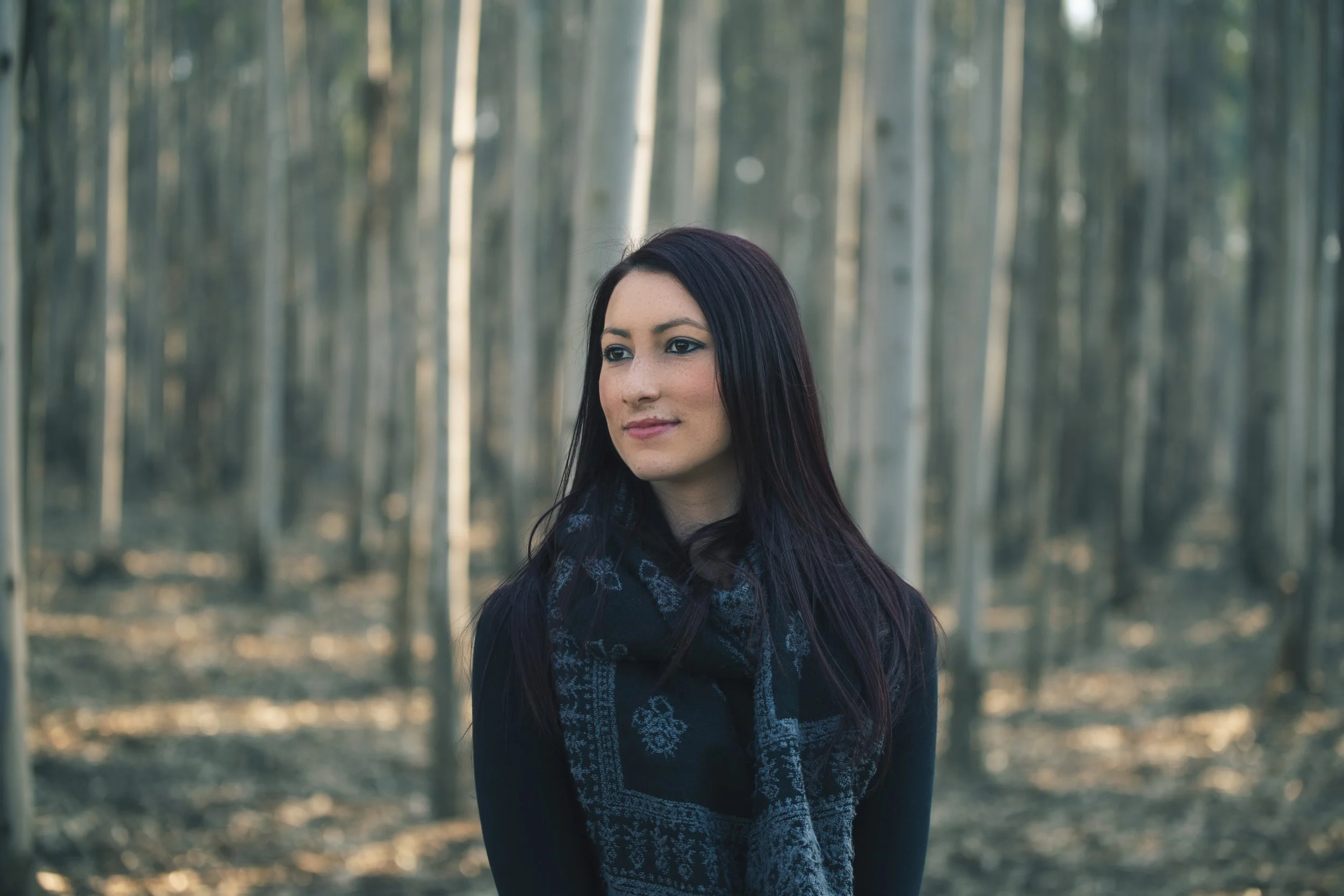 A woman with long dark hair standing in a forest with tall, thin trees. She is wearing a dark top and a patterned scarf, looking slightly to the side.