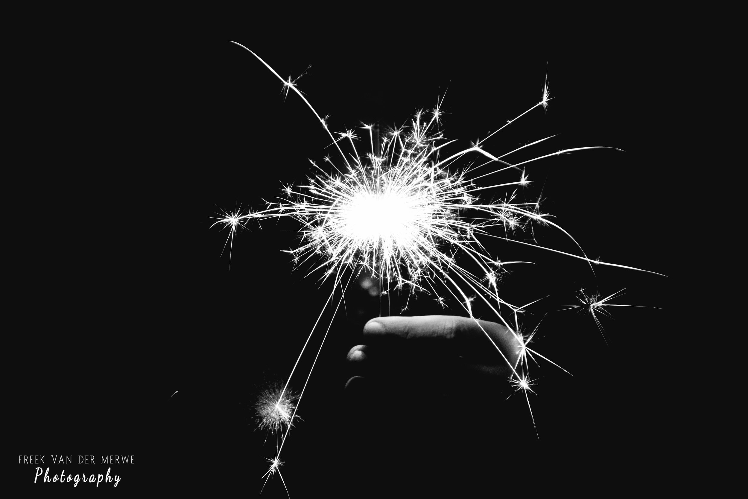 A hand holding a lit sparkler with bright sparks against a dark background.