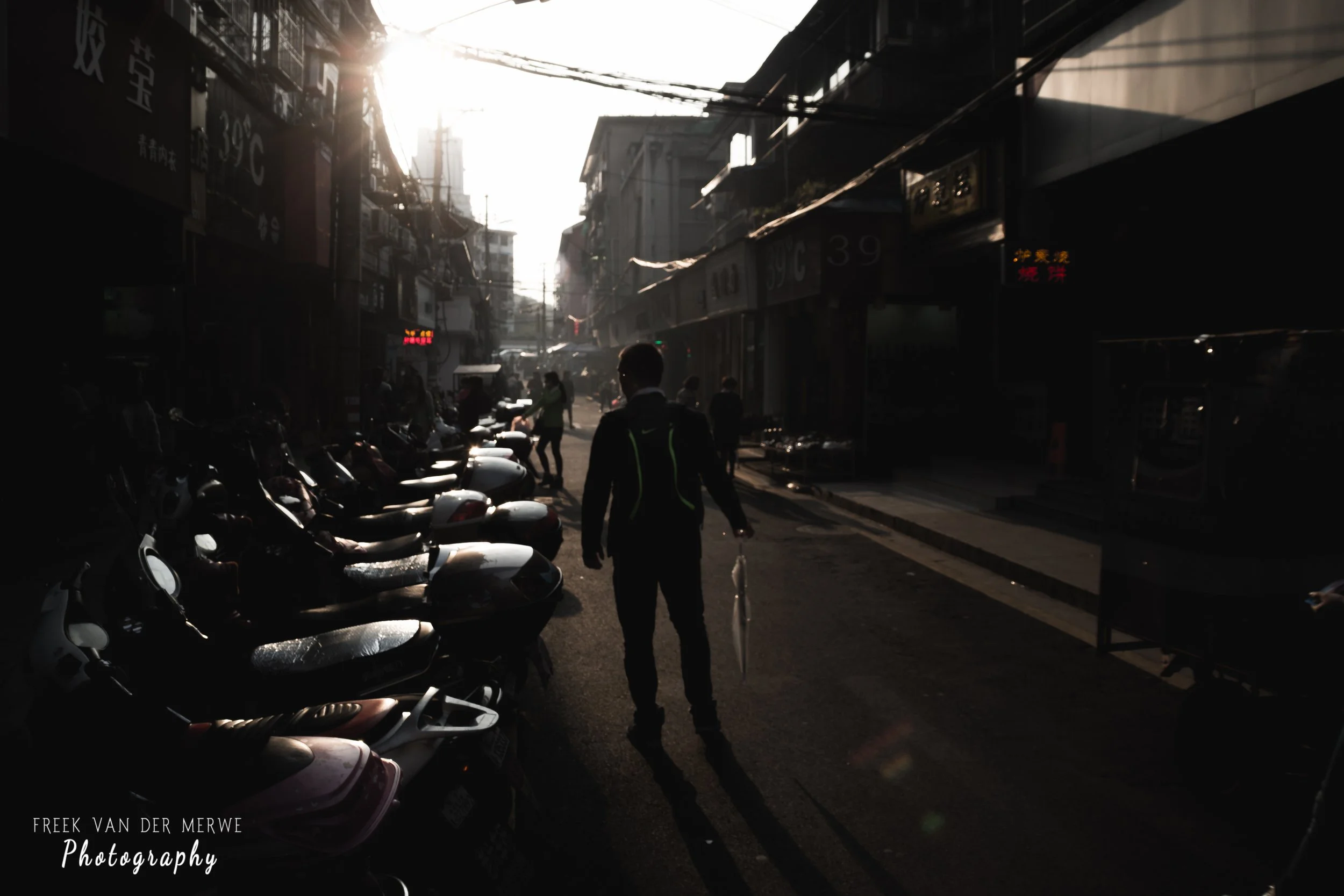 A man walking down a narrow street lined with parked scooters and small shops, with the sun setting in the background casting long shadows.