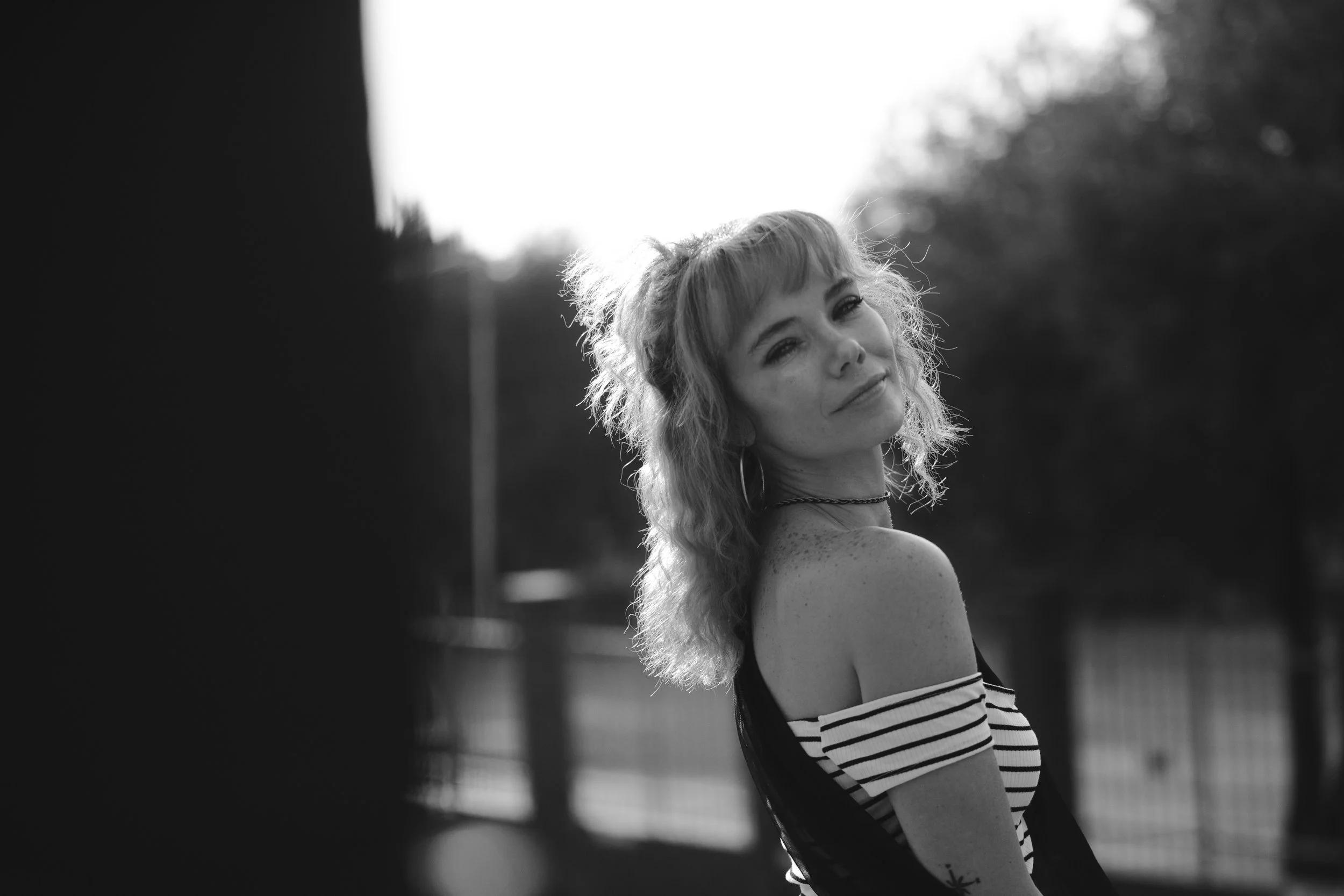 Black and white photo of a young woman with wavy hair, wearing a striped off-the-shoulder top, looking over her shoulder with a slight smile outdoors.