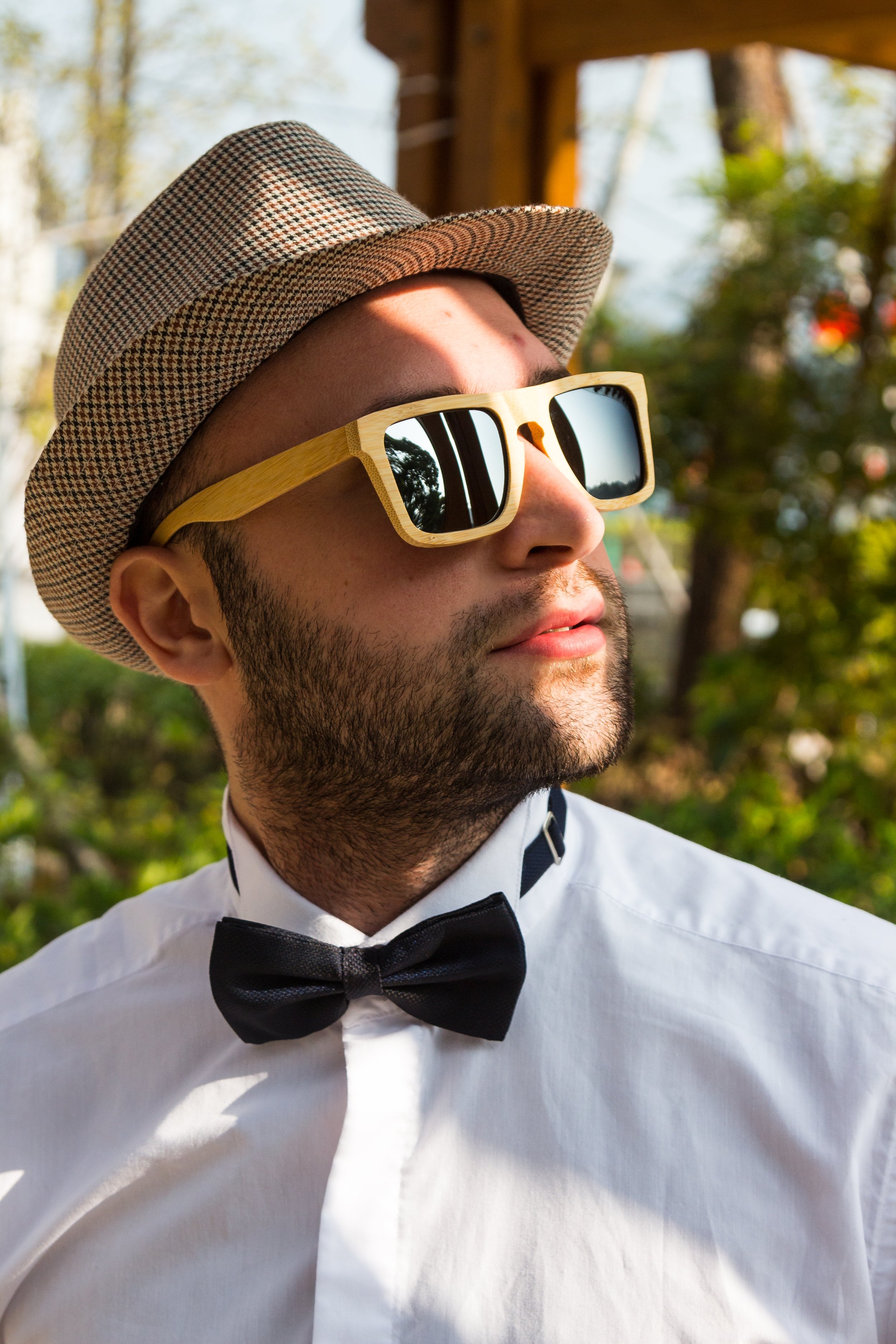 A man wearing a patterned fedora, large wooden sunglasses, a white dress shirt, and a black bow tie outdoors during daytime.