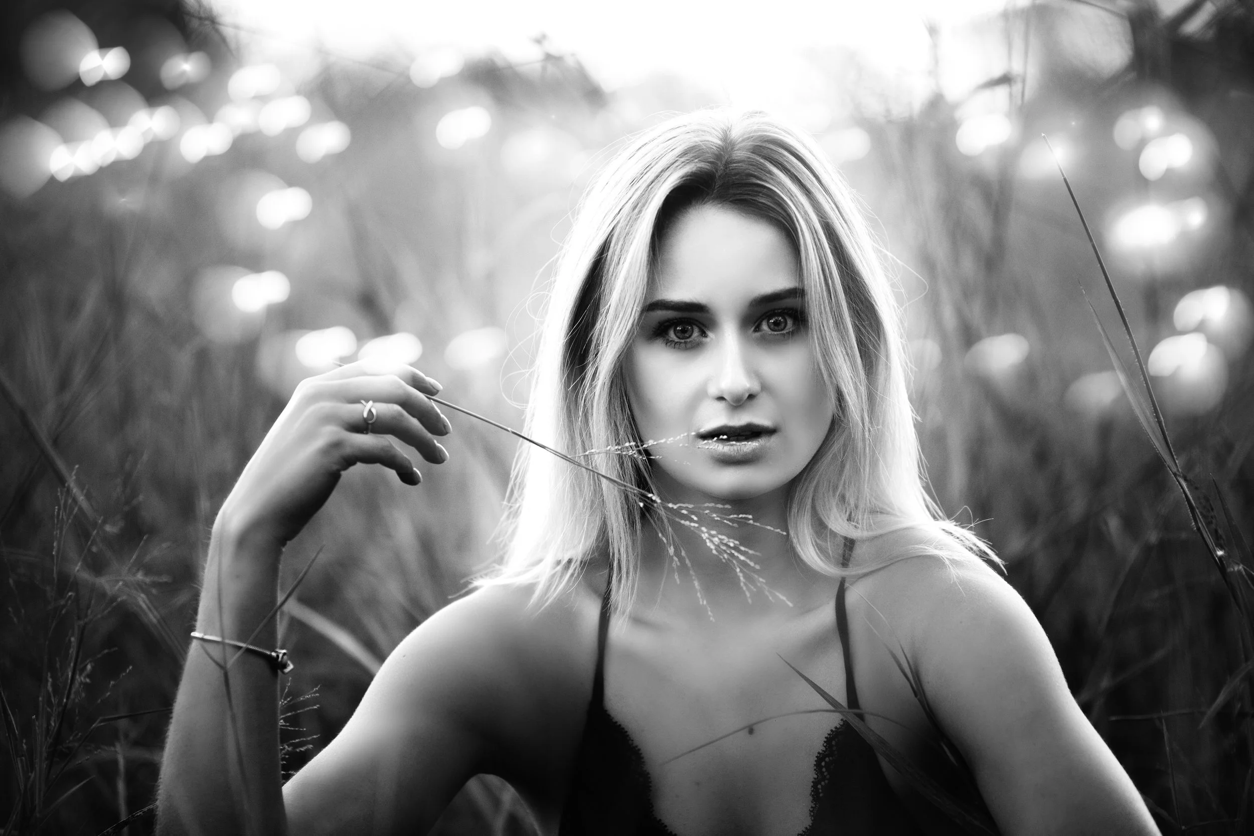 A black and white photo of a young woman with blonde hair in a field, holding a piece of grass near her face and looking directly at the camera.
