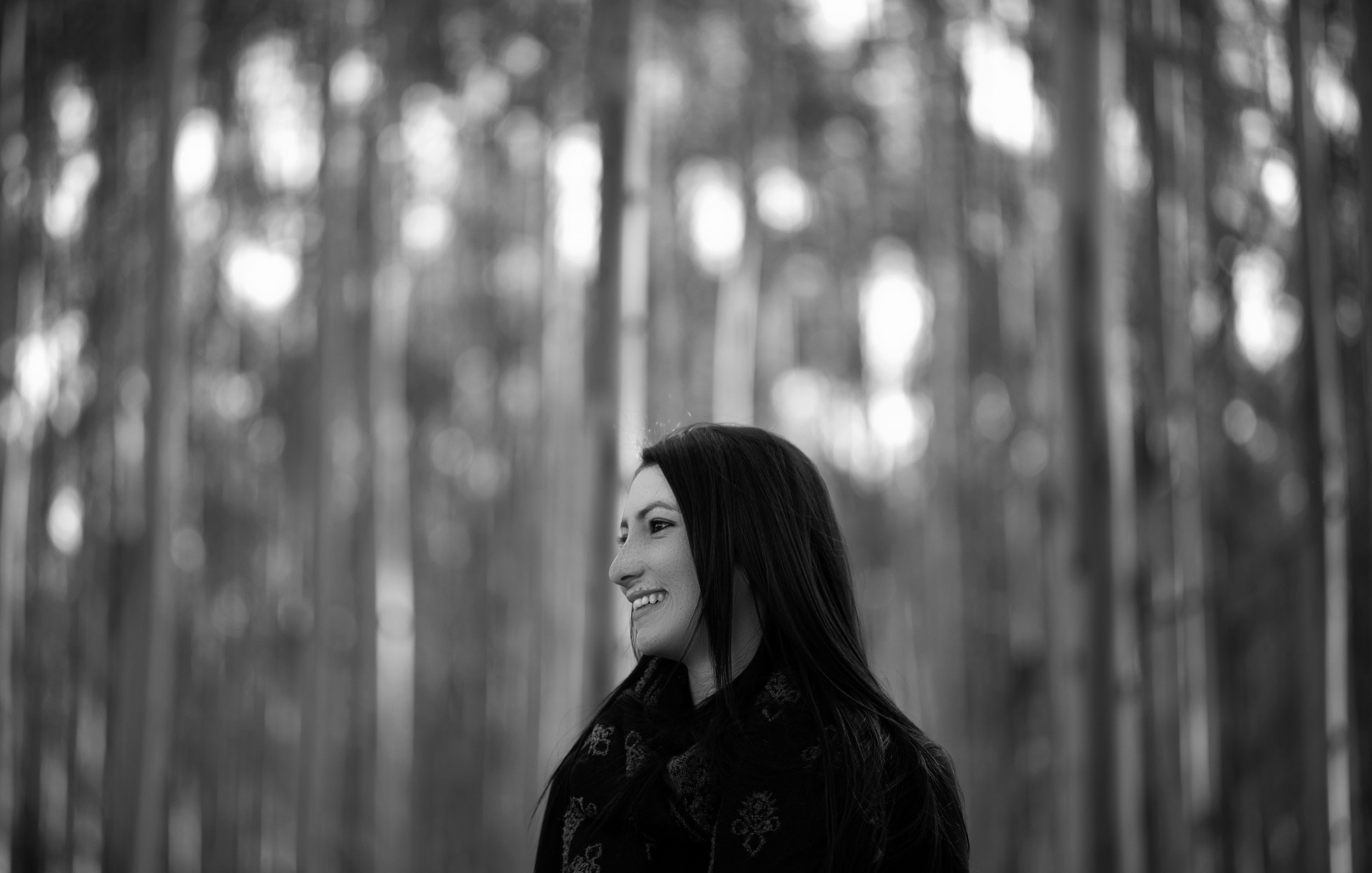 A woman with long dark hair smiling in a forest with tall trees, black and white photograph.