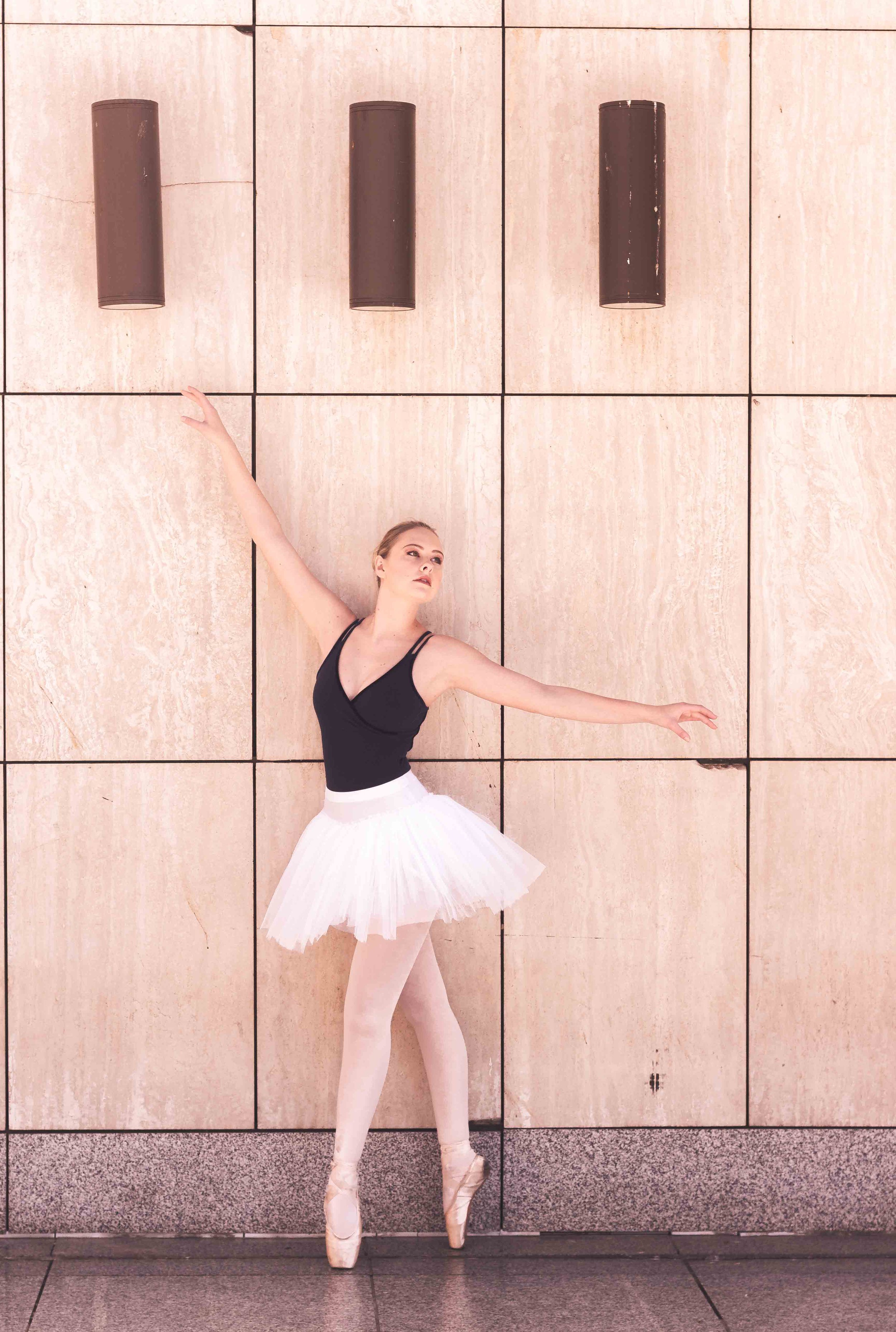 Ballet dancer in a black leotard and white tutu practicing ballet on pointe in front of a tiled wall.