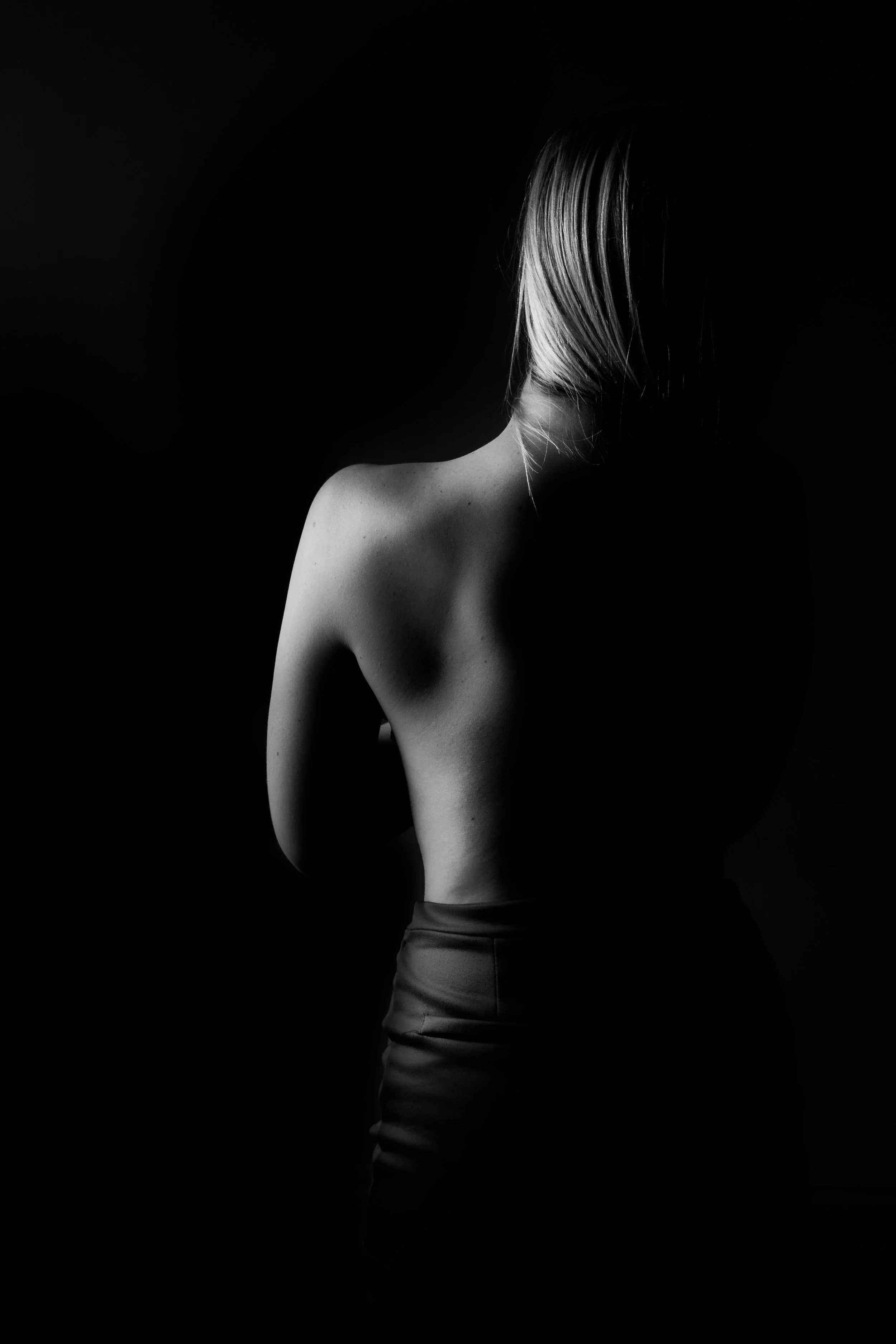 Black and white photo of a woman with bare back and shoulder, facing away, with her face turned slightly down, hair covering part of her face, in a dark background.