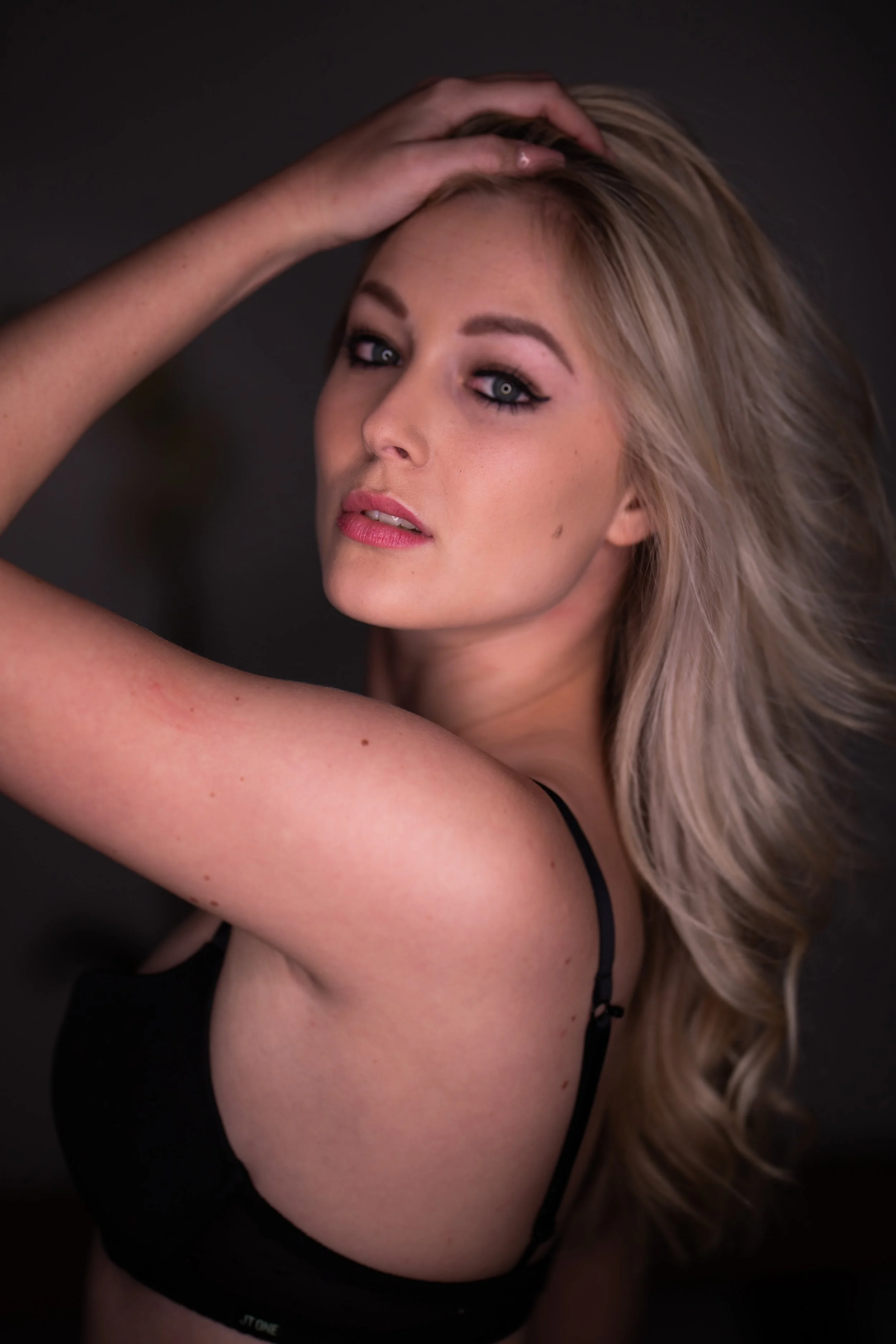 A woman with blonde hair and makeup posing with her hand on her head, wearing a black bra.