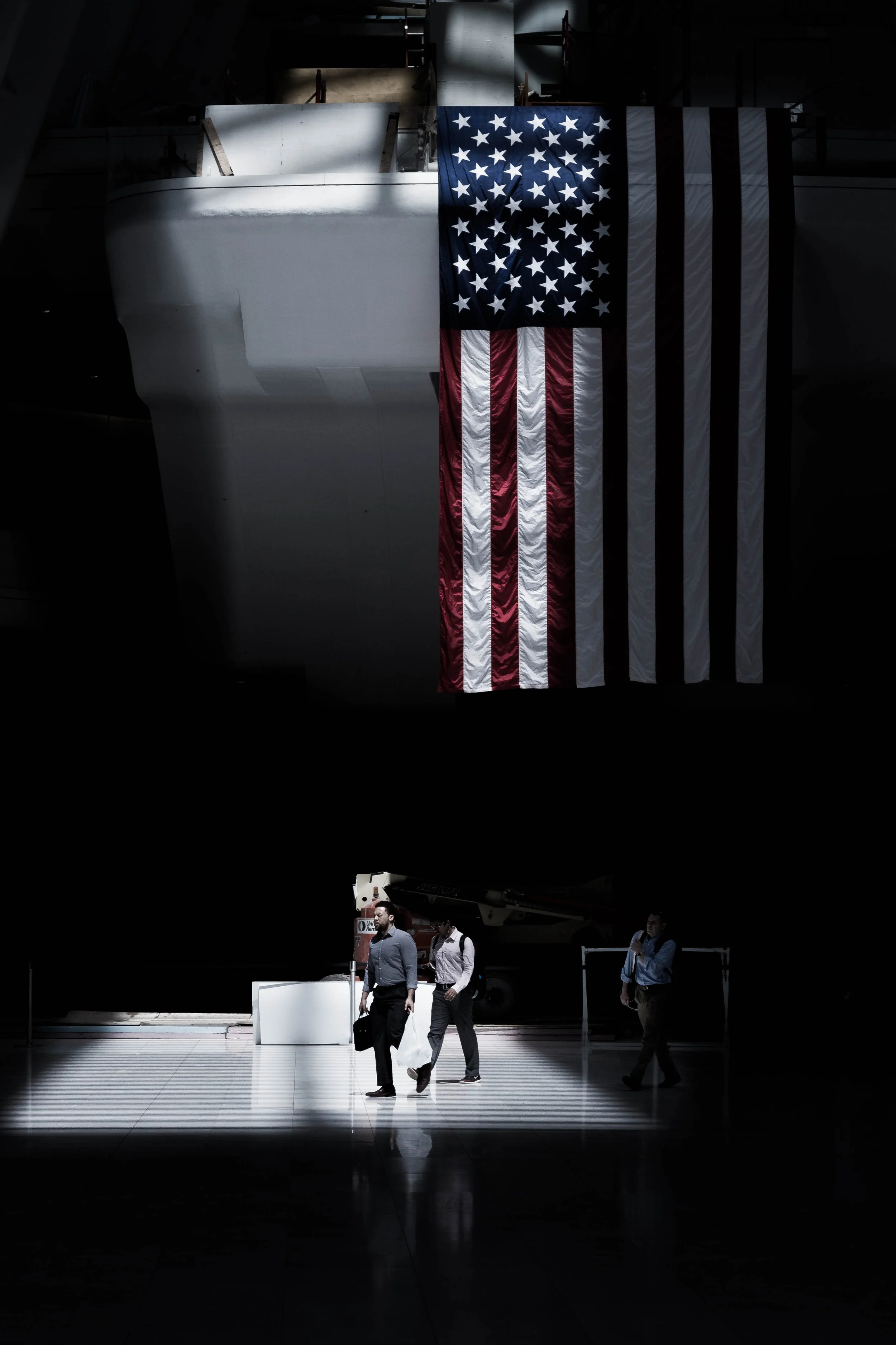 An American flag hanging inside a dark airport or exhibition hall with three business people walking in the foreground.