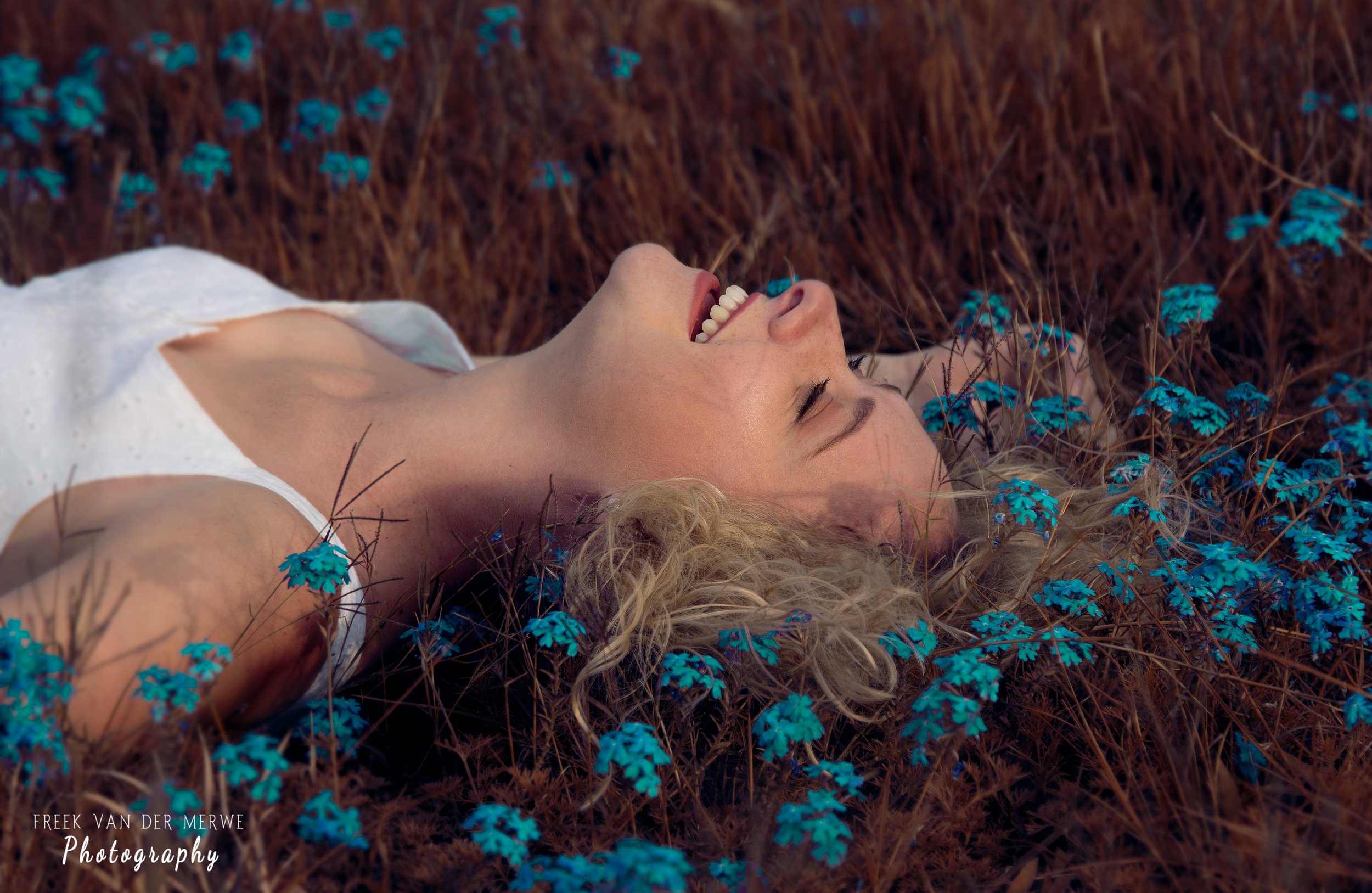 A woman with curly blonde hair lying on her back in a field of brown grass and small blue flowers, smiling with her eyes closed.