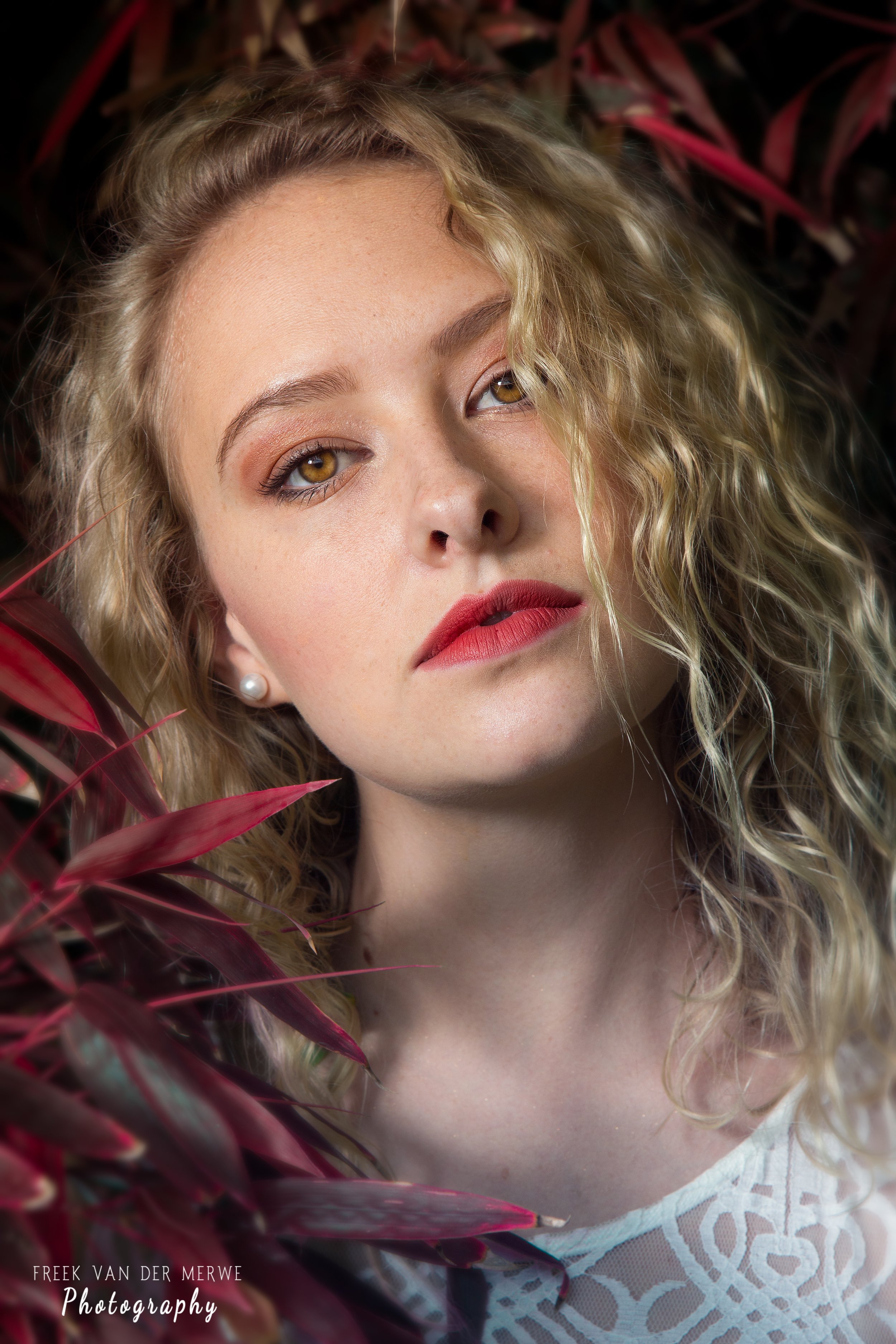 Close-up portrait of a young woman with curly blonde hair, wearing pearl earrings and red lipstick, surrounded by red and purple leaves.
