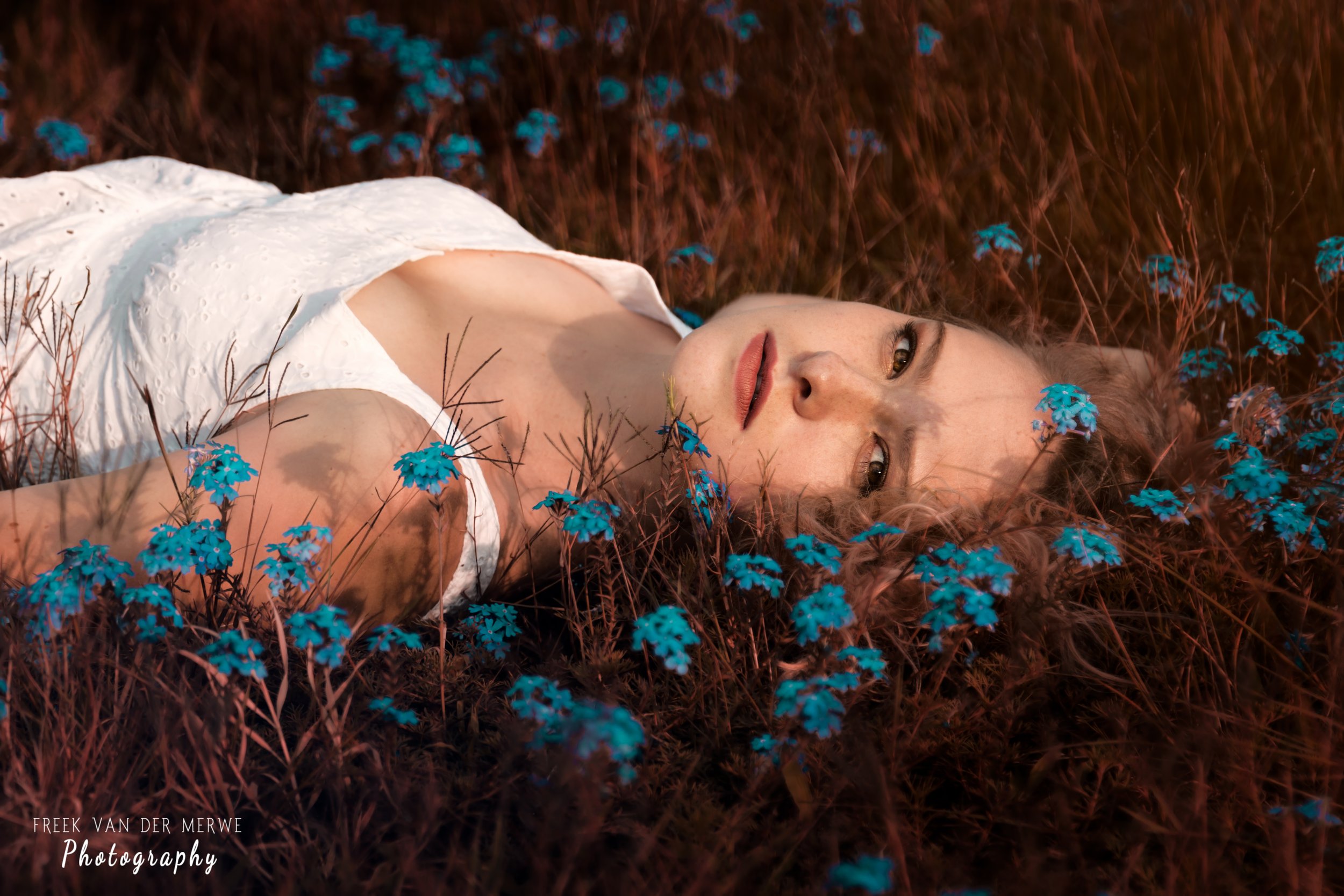 A woman in a white dress lying on the ground covered with small blue flowers and dry grass, looking at the camera with a relaxed expression.