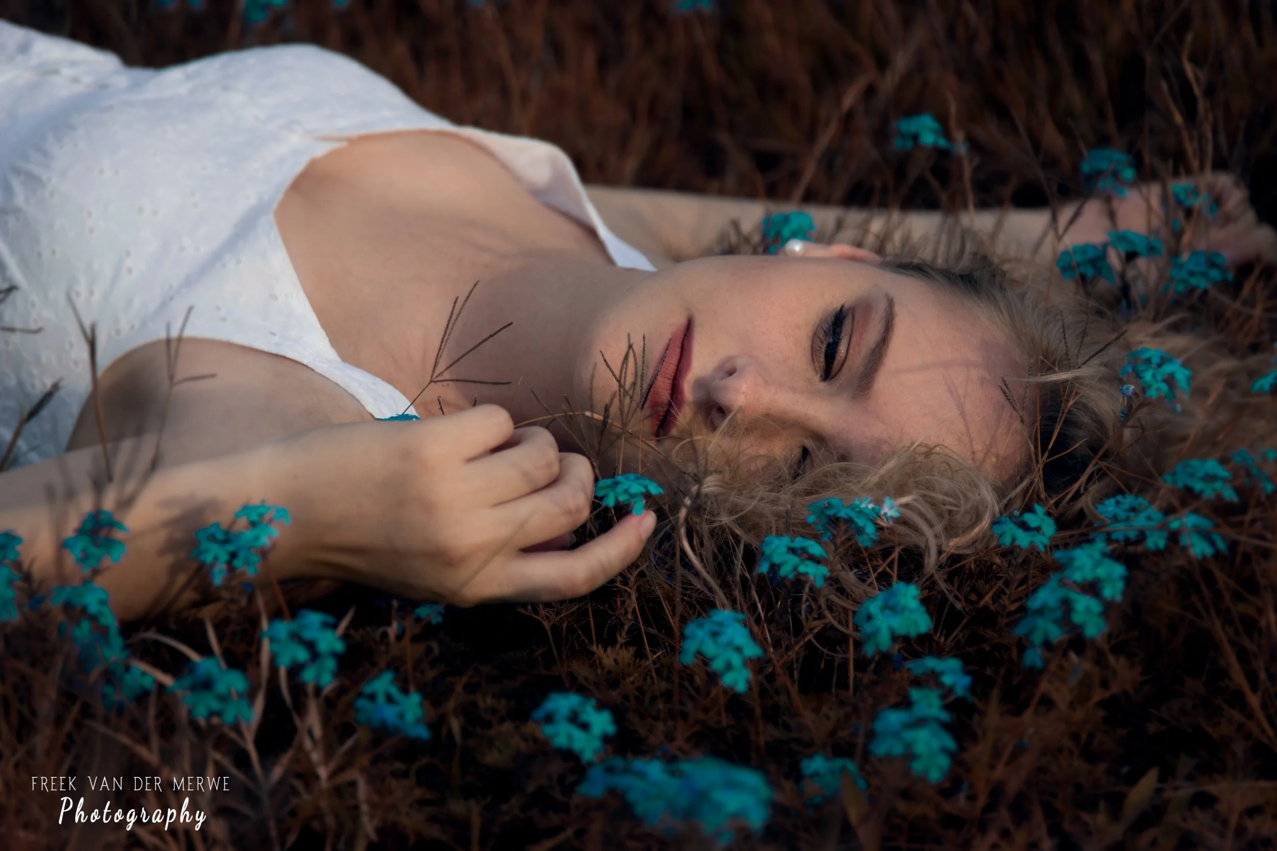 A woman lying on the ground with brown grass and small blue flowers around her, wearing a white top, with her eyes closed and her hand holding a blue flower.