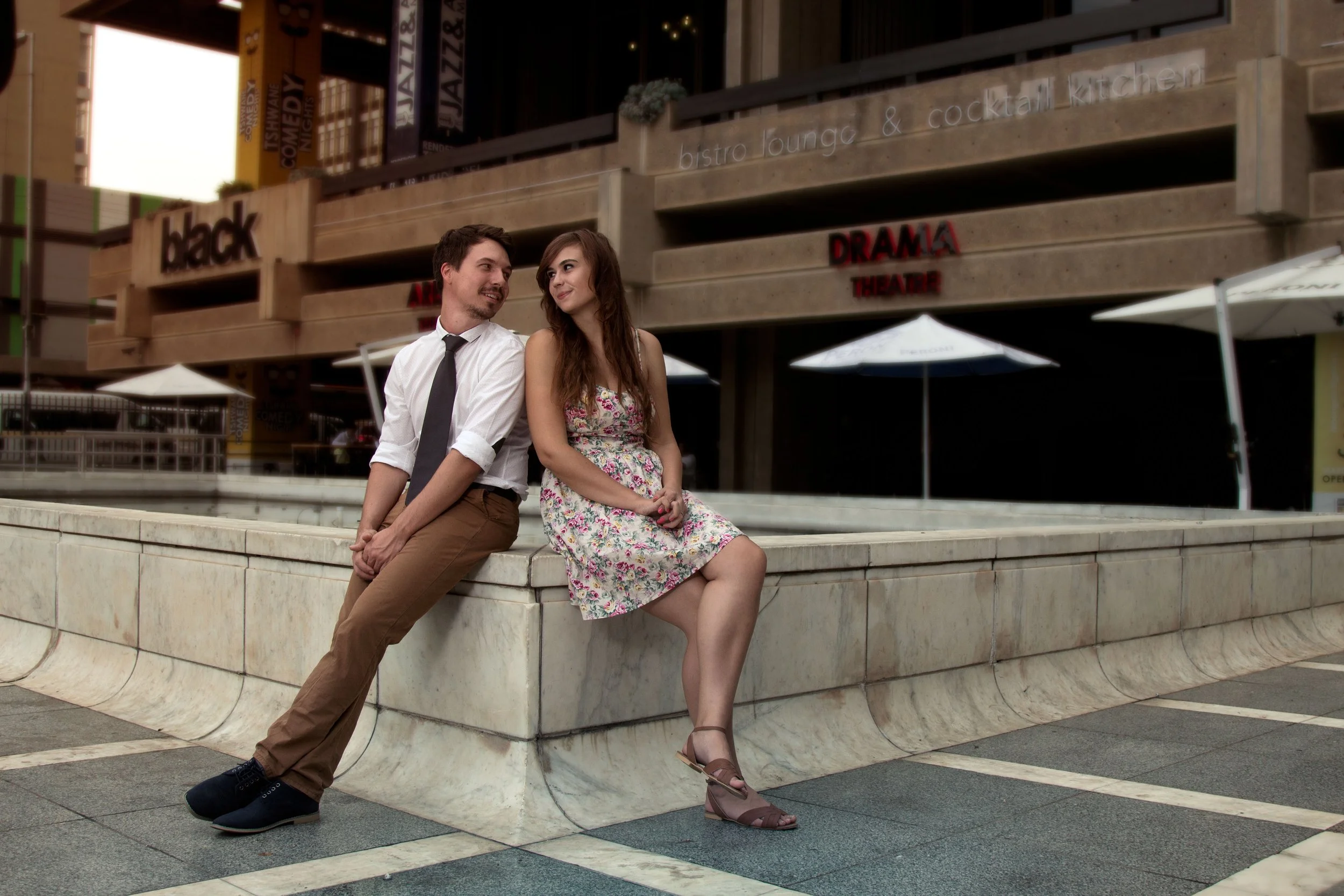 A young man and woman sitting on a marble ledge outside a building with signs for a drama theater and a bistro lounge, engaged in conversation and smiling at each other.