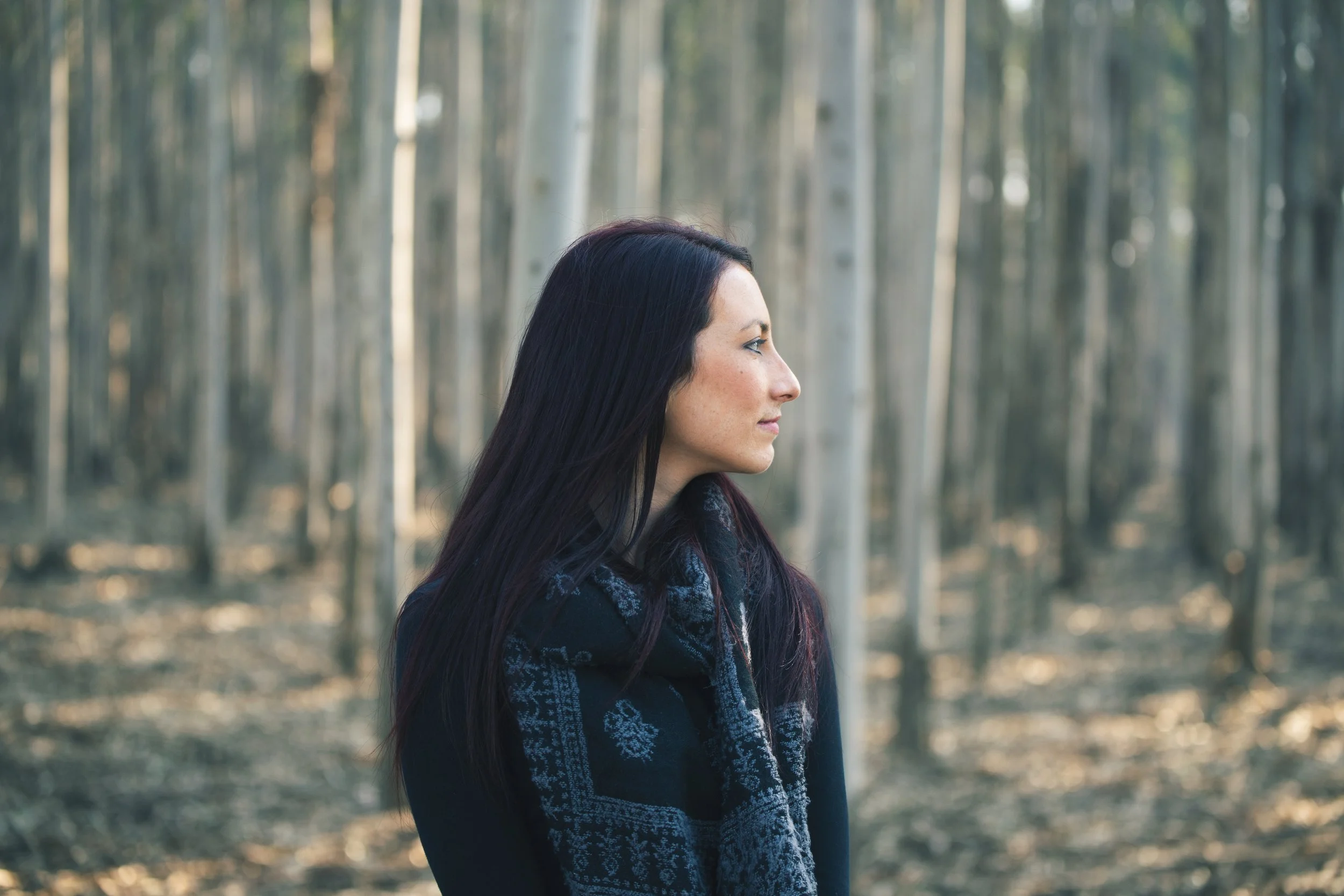 A woman with long dark hair standing in a forest of tall, thin trees with light-colored bark, wearing a dark jacket and a patterned scarf.