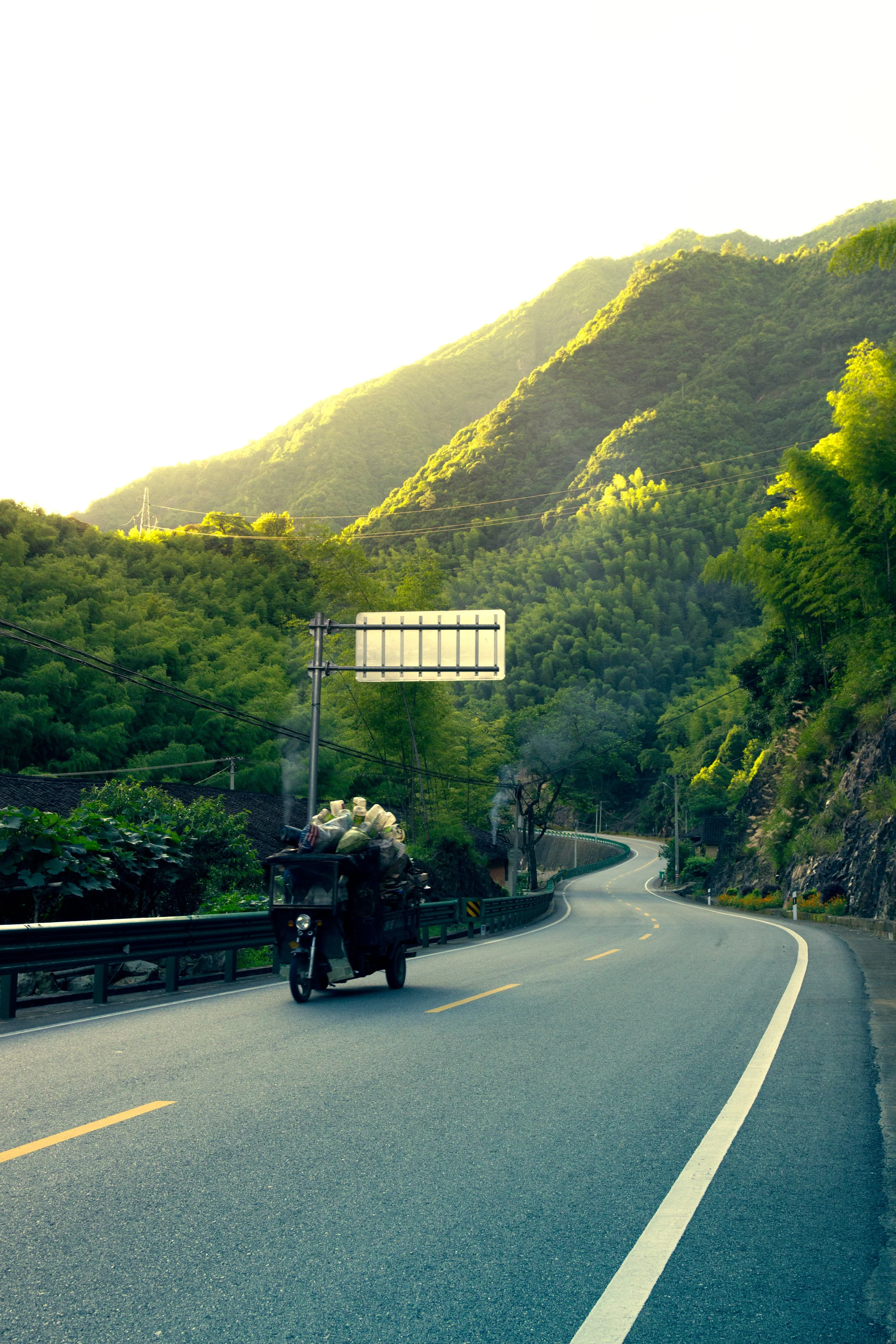 Curving mountain road with a motorcycle carrying cargo, surrounded by green forests and hills under a bright sky.