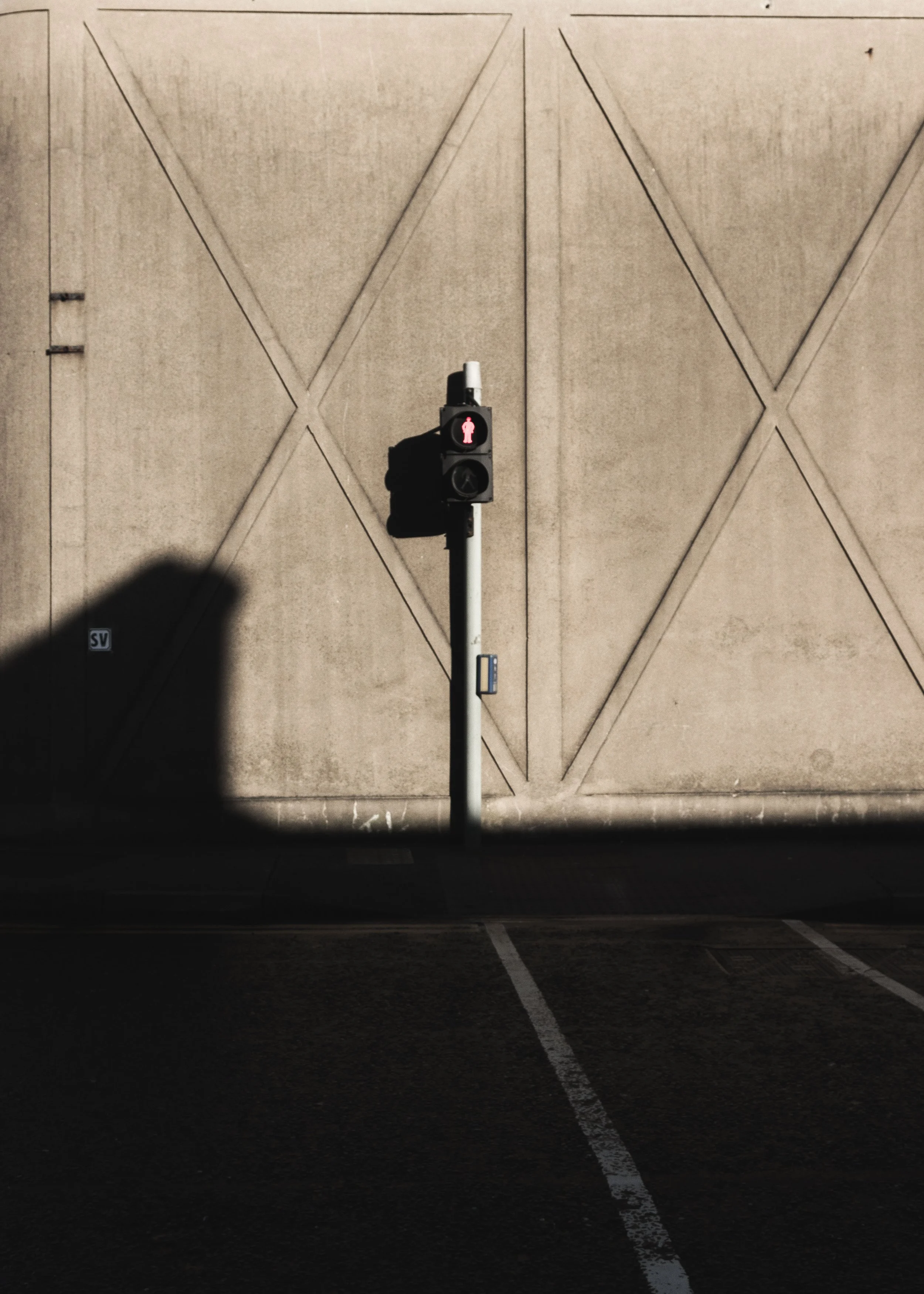 A pedestrian traffic light with a red figure, mounted on a white pole in front of a plain beige wall with geometric line patterns. The shadow of a person is cast on the wall.