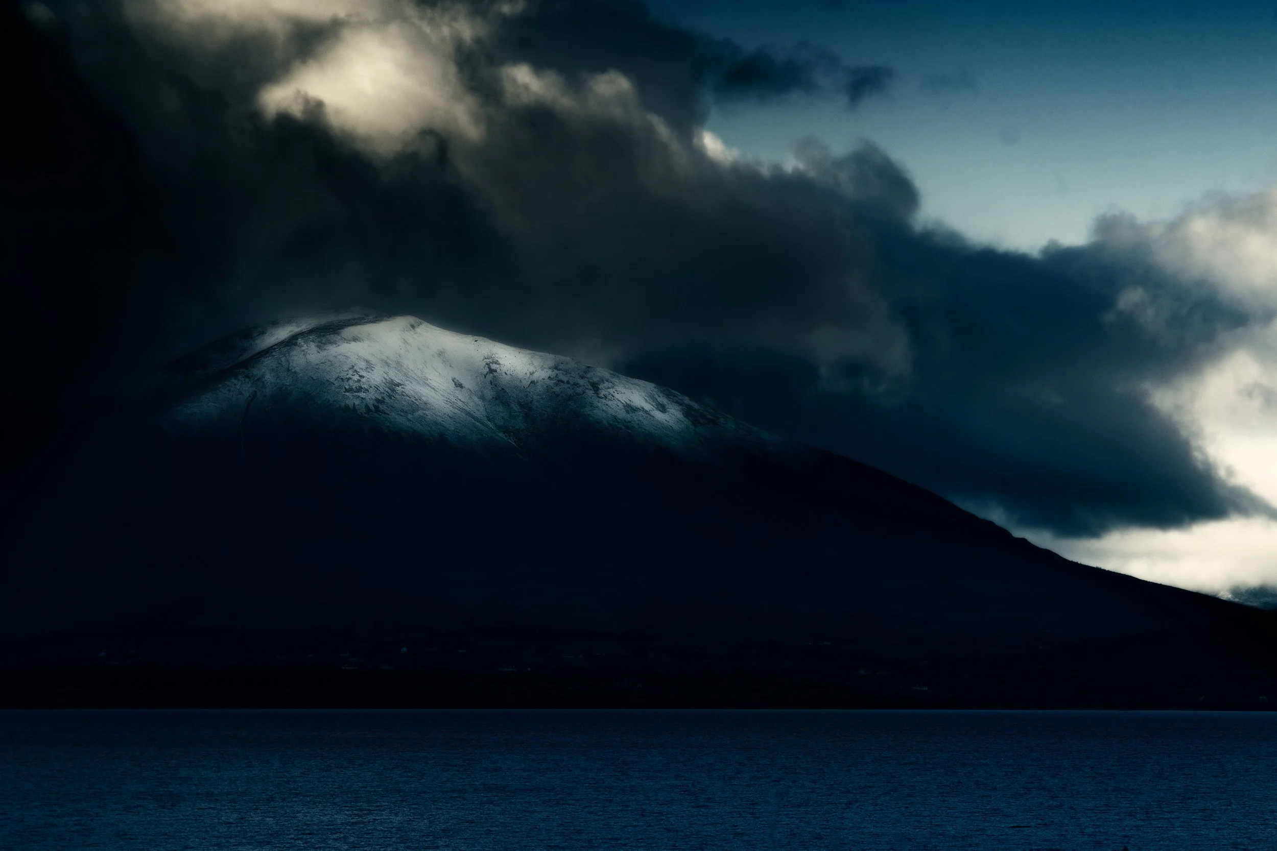 A snow-capped mountain partially obscured by dark clouds over a body of water.