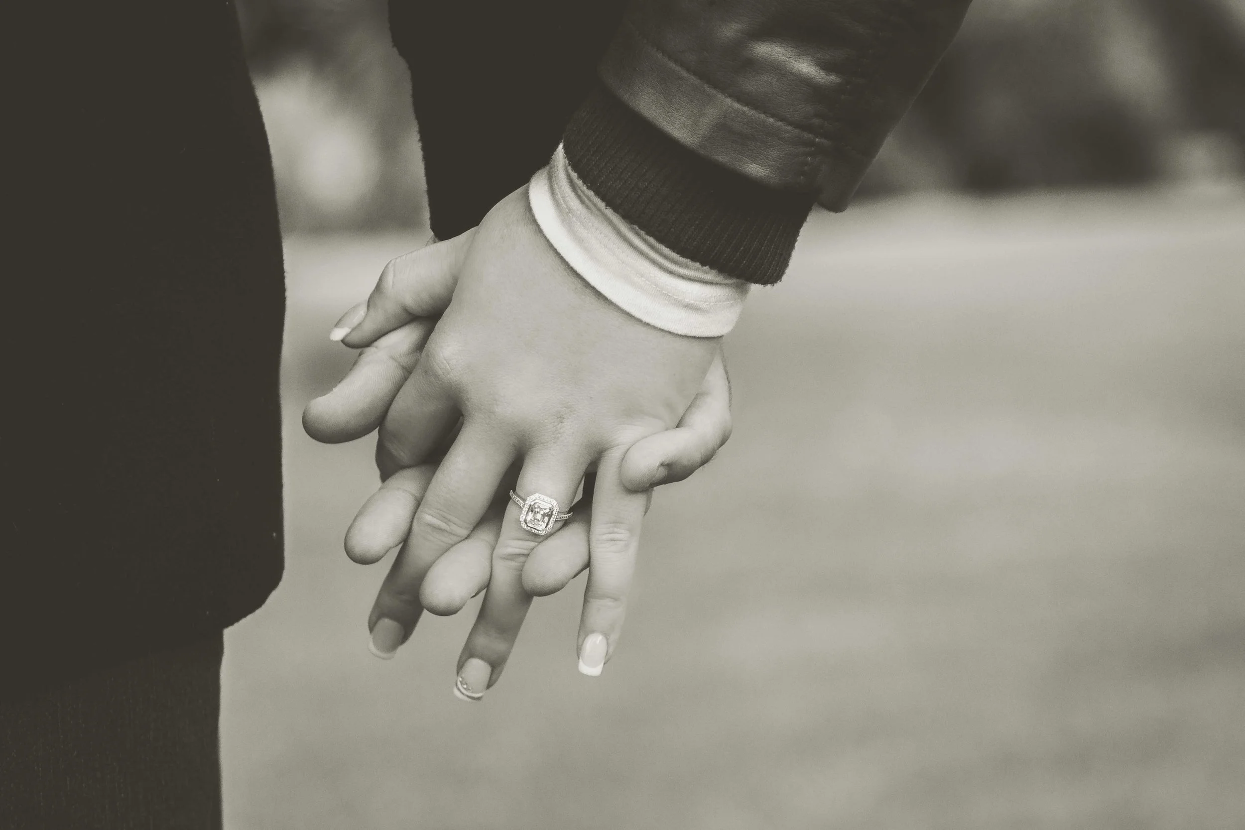 Close-up of a couple holding hands, with one person's hand resting on top of the other's. The person on top wears a ring on their finger, and both are wearing jackets, with the cuff of the sleeve visible. The background is blurred.