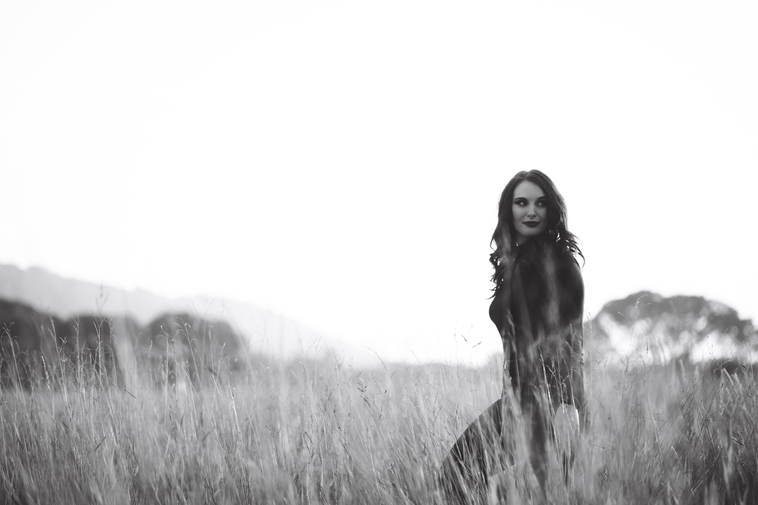 A black and white photo of a woman standing in a grassy field, looking over her shoulder with a slight smile.