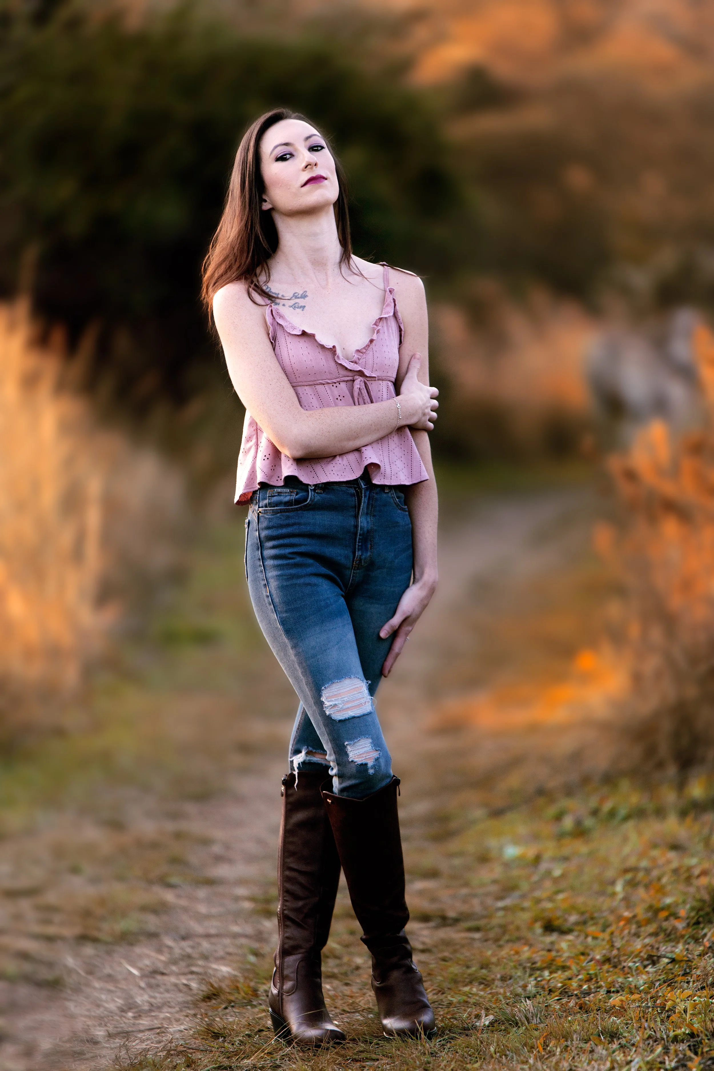 A young woman with long brown hair and fair skin standing outdoors on a dirt path, wearing a pink sleeveless ruffled top, ripped jeans, and black knee-high boots, with her arms crossed and looking confidently at the camera.