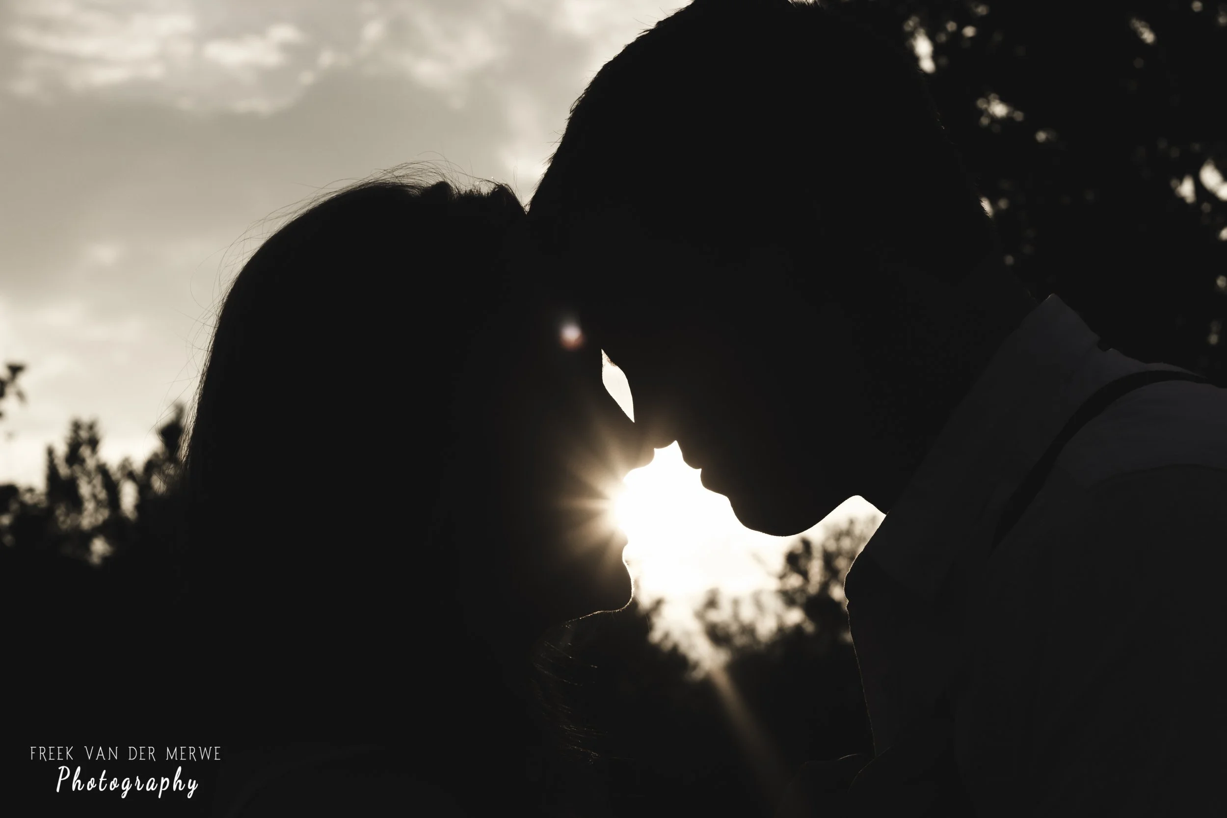 Silhouettes of a couple with foreheads and noses touching, backlit by the setting sun, with trees and clouds in the background.