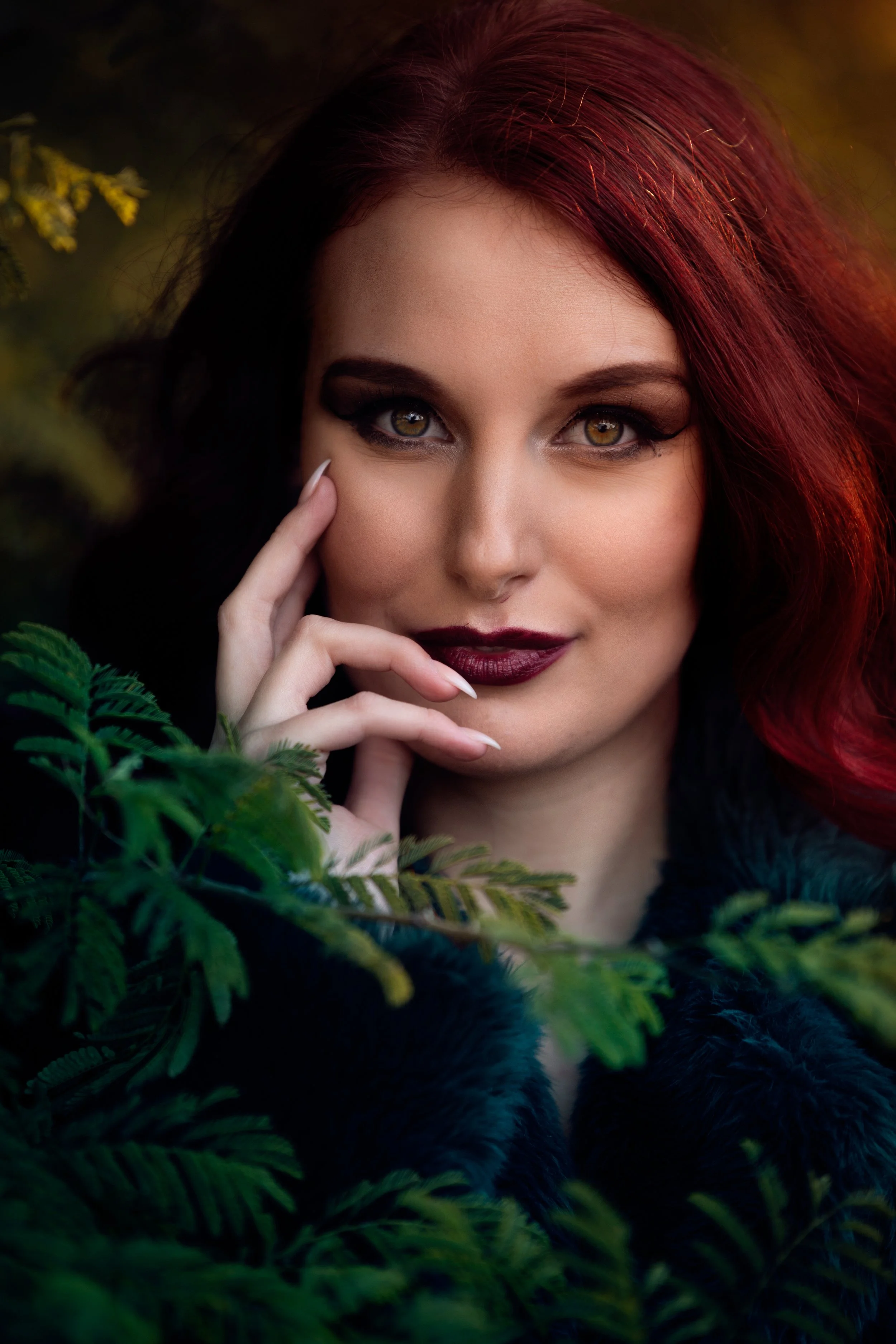 A woman with red hair and dark makeup among green foliage, looking directly at the camera.