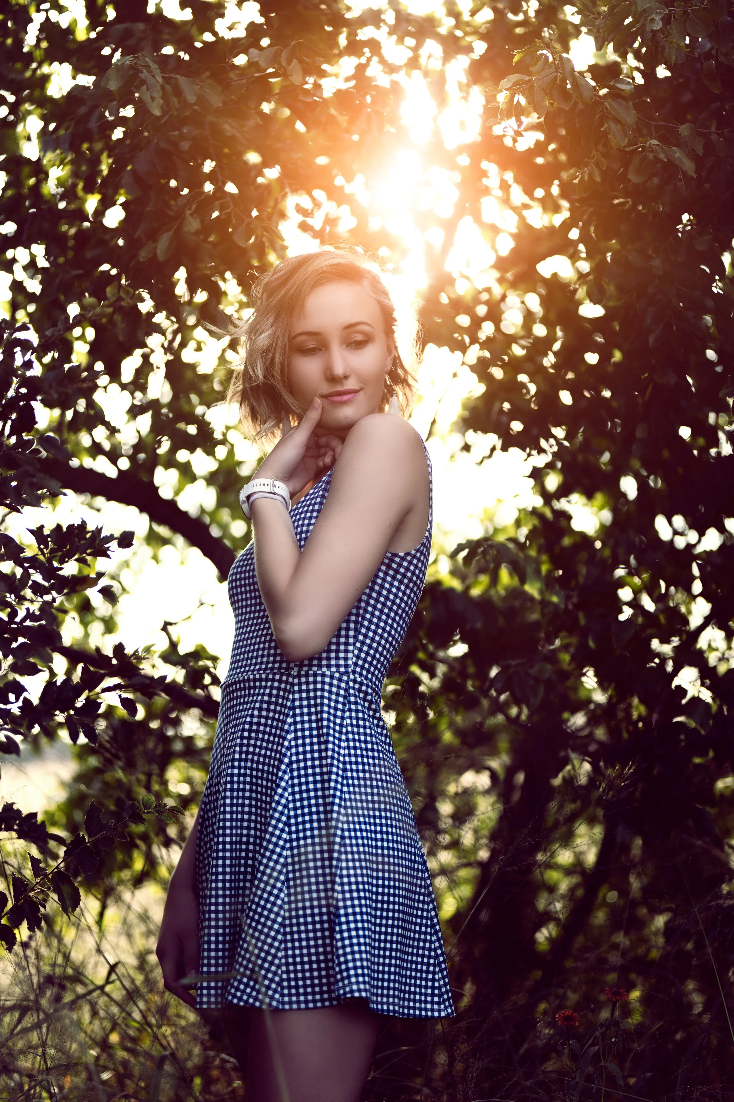 A young woman with short, wavy hair standing outdoors among trees during sunset. She is wearing a blue and white checkered dress and a white wristwatch, with her eyes closed and her hand resting gently on her neck, as sunlight filters through the lea