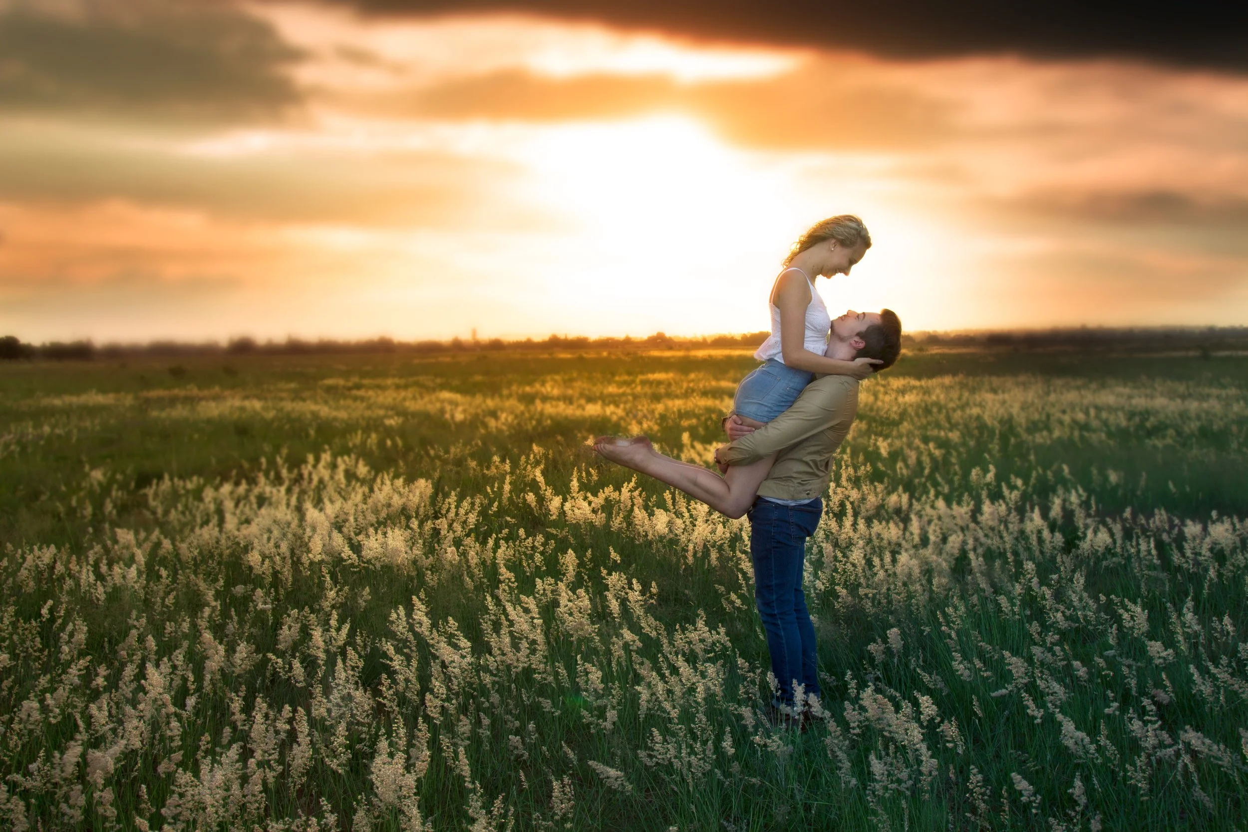 A man lifting a woman in a field of tall grass and flowers at sunset