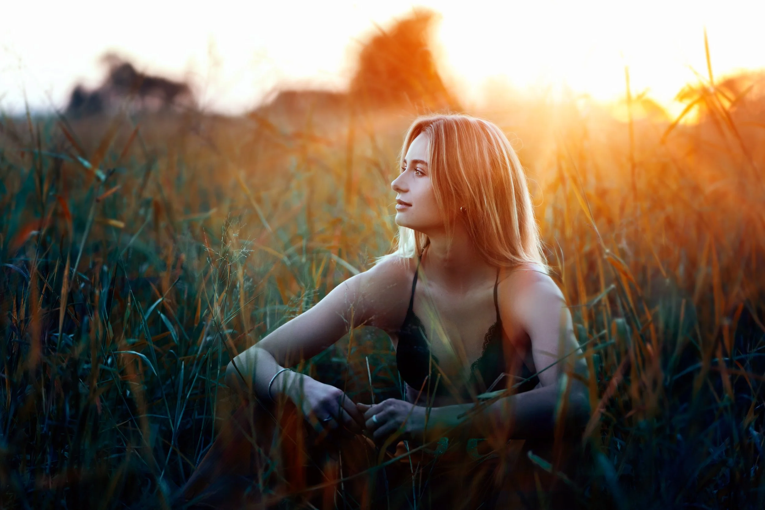 A woman with blonde hair sitting in a grassy field during sunset, looking to the side.