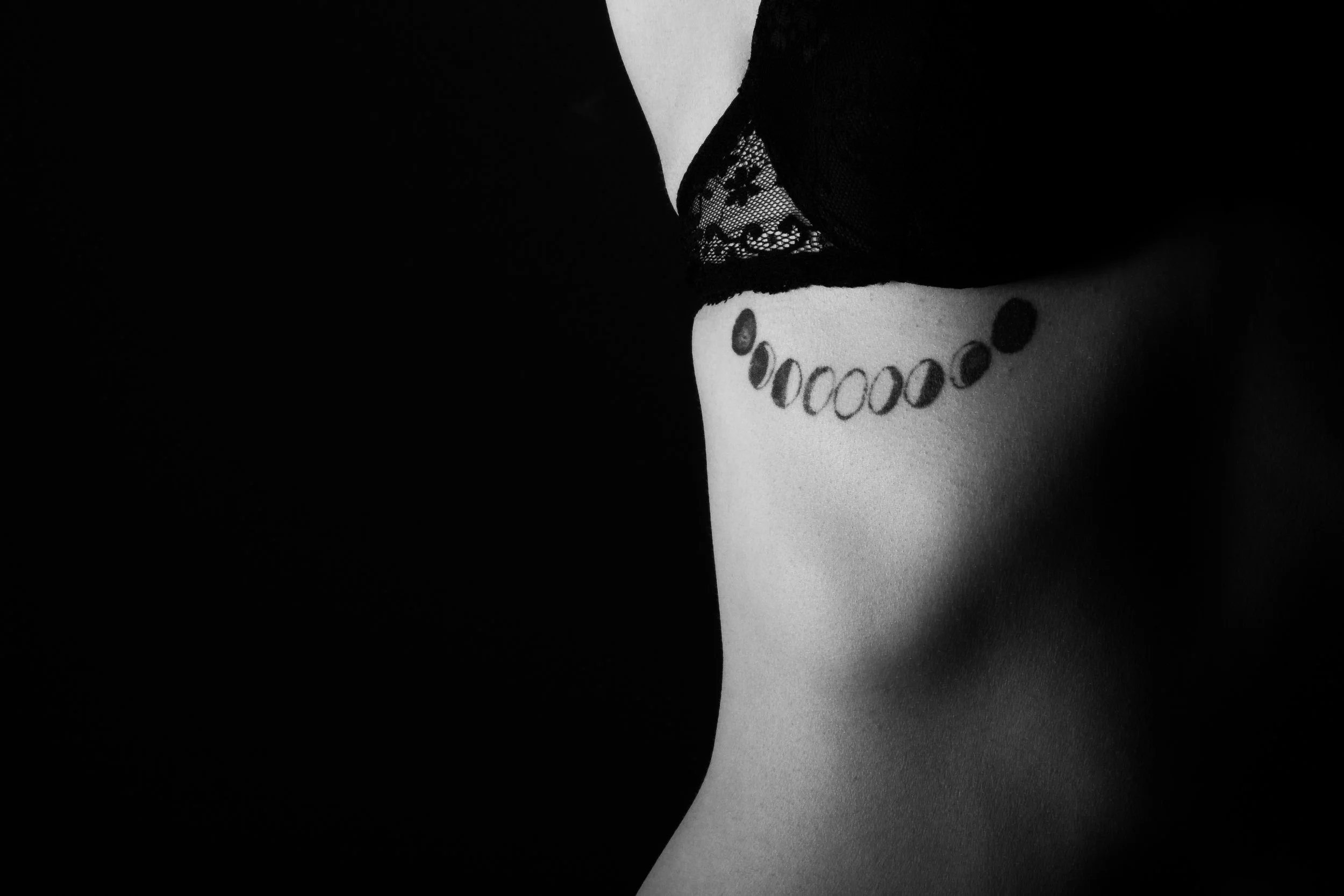 Close-up of a tattoo of a moon phases design on a person's side ribcage, with lace clothing visible at the top.