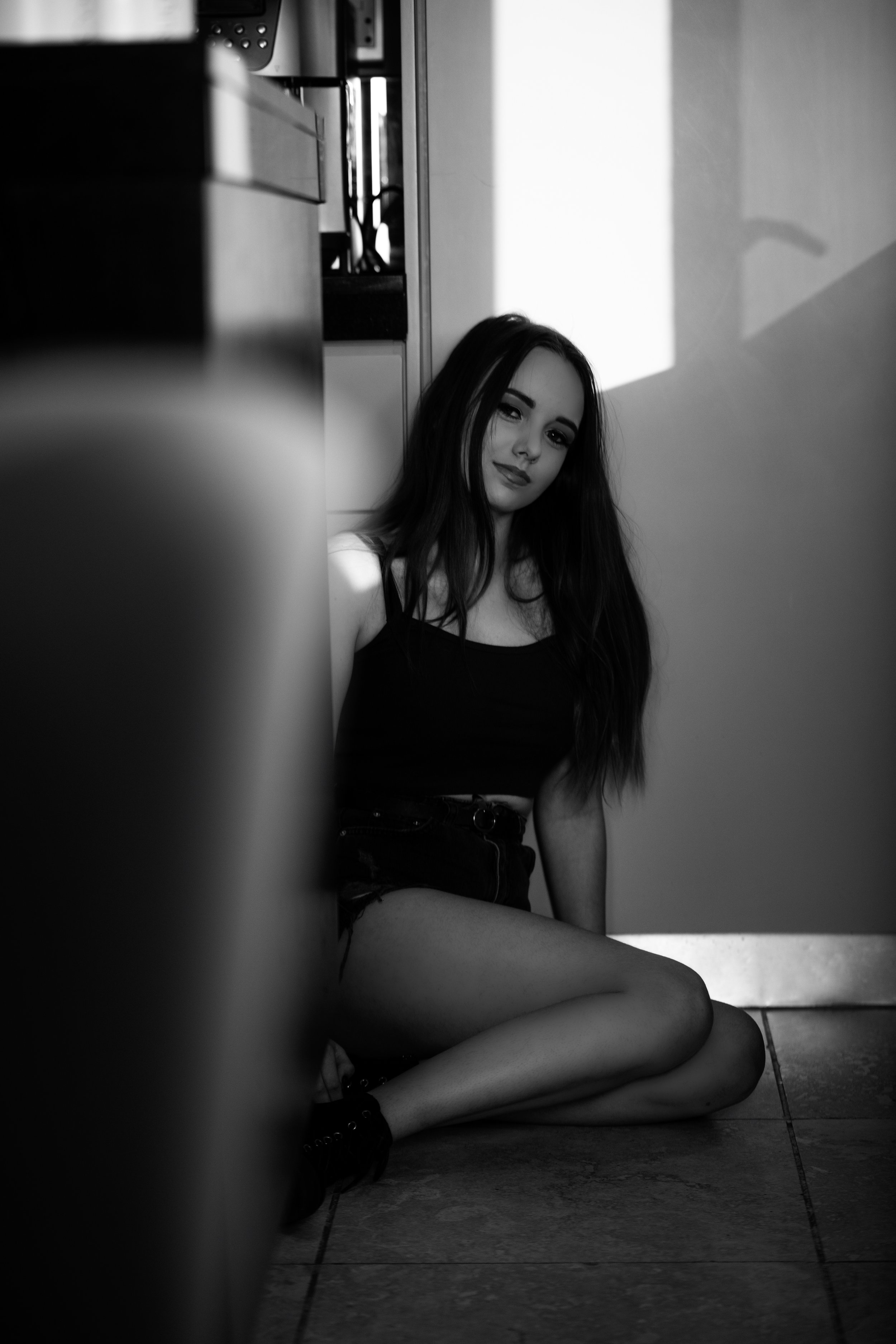 A young woman with long dark hair sitting on the floor, leaning against a wall, in a room with light falling on her face and body. The photo is in black and white.