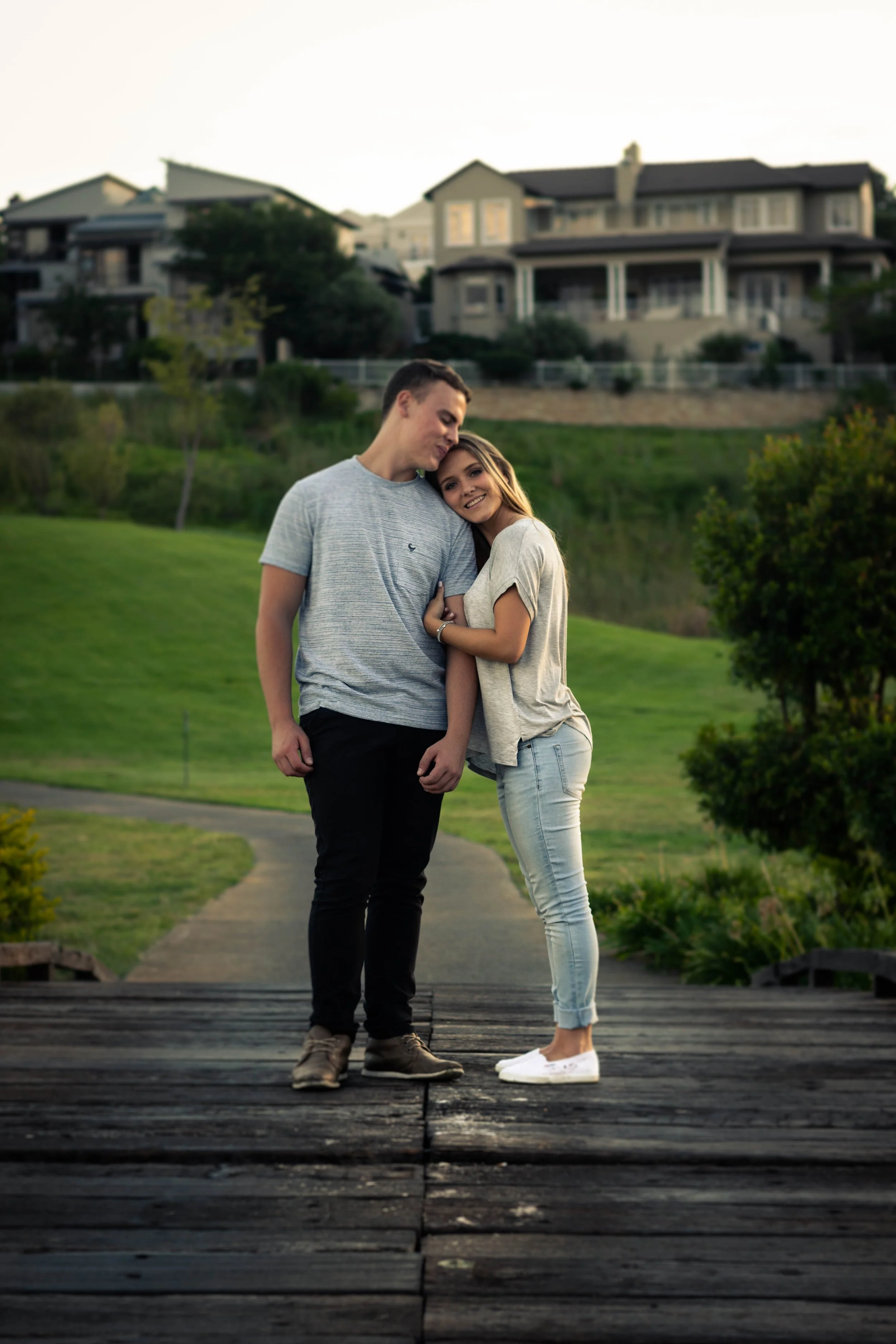 A young couple stands on a wooden bridge in a park, embracing each other with happy expressions, with greenery and houses in the background during twilight.
