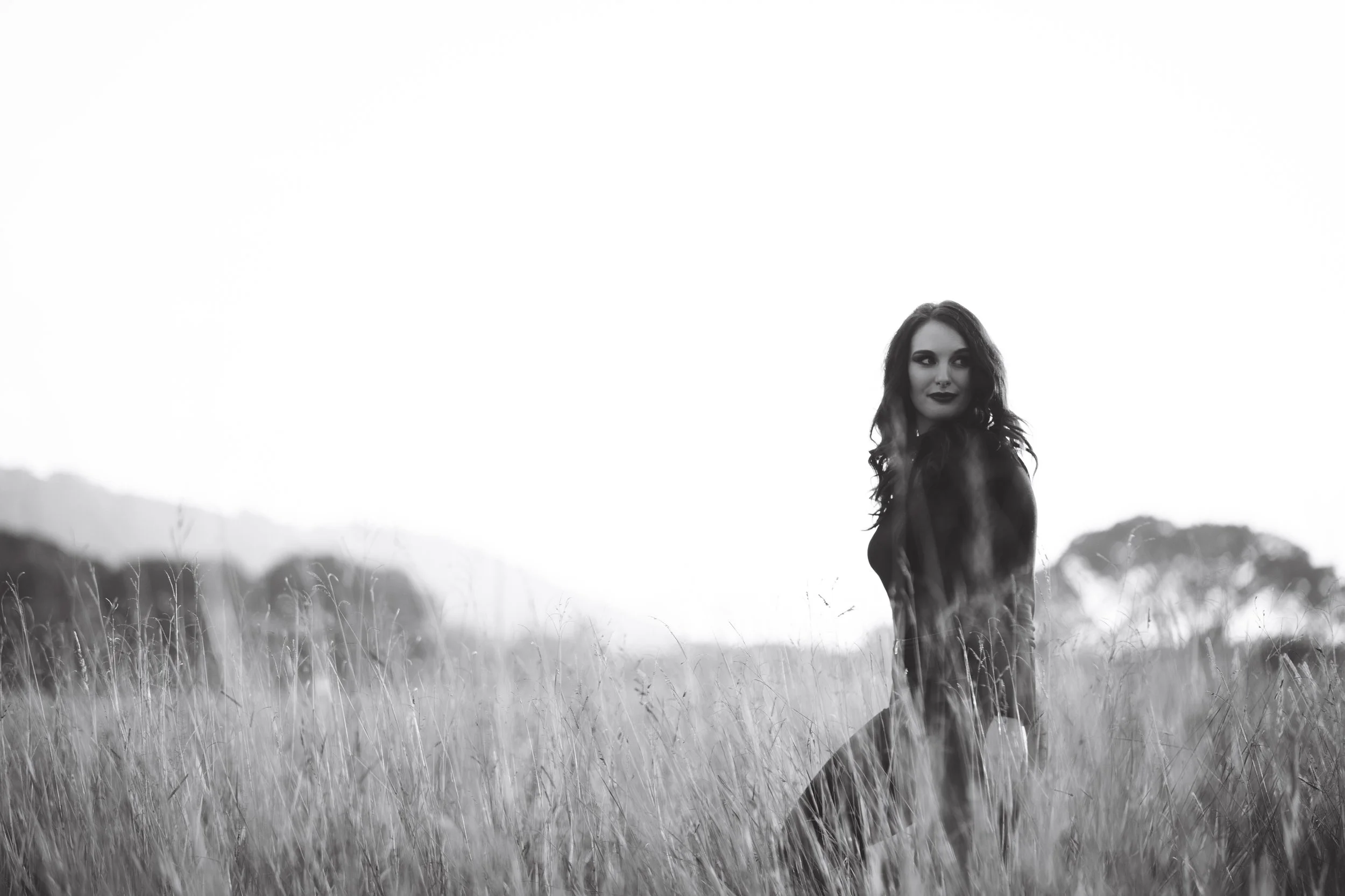 A woman with dark hair and makeup standing in a field of tall grass with mountains and trees in the background, black and white photo