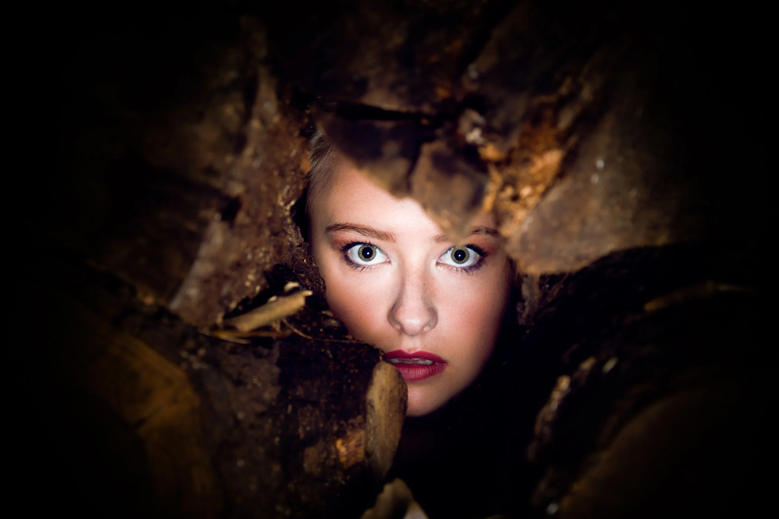 A young woman's face peering through a hole in stacked wood logs, with bright blue eyes and red lipstick.