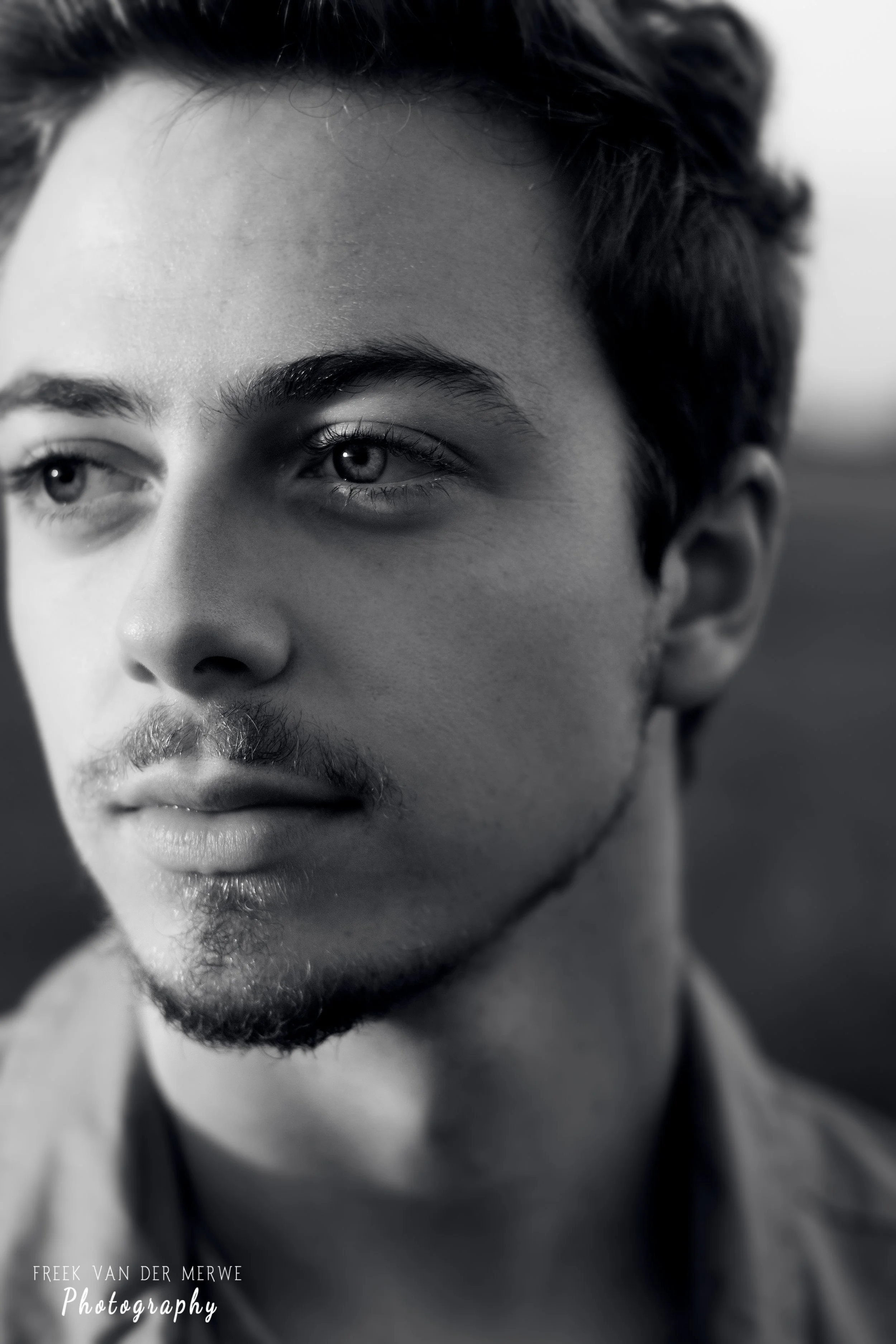 Close-up of a young man with light skin, short wavy hair, and facial hair, looking to the side with a thoughtful expression in black and white.
